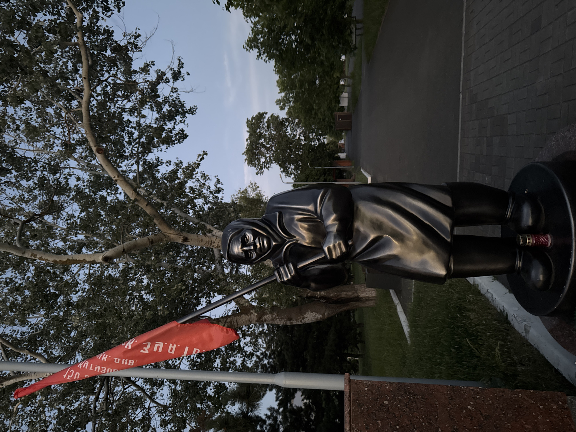 This is a full-body, eye-level, outdoor, vertical photograph of a dark-colored metal statue of a woman holding a red flag. The statue is positioned in front of a walkway lined with trees and a grassy area. The statue depicts a woman wearing a headscarf and a long coat, with her hands clasped around a flag pole. The flag is red with white Cyrillic text. A single red candle in a glass holder is placed at the base of the statue. To the left of the statue is a portion of a red granite monument. The walkway behind the statue is paved with dark asphalt, and there is a brick-like pattern on the ground to the right of the walkway. The sky is overcast and a light blue color, indicating either dawn or dusk. The image captures a somber and memorial atmosphere. This statue is known to be located in Mariupol, Ukraine.