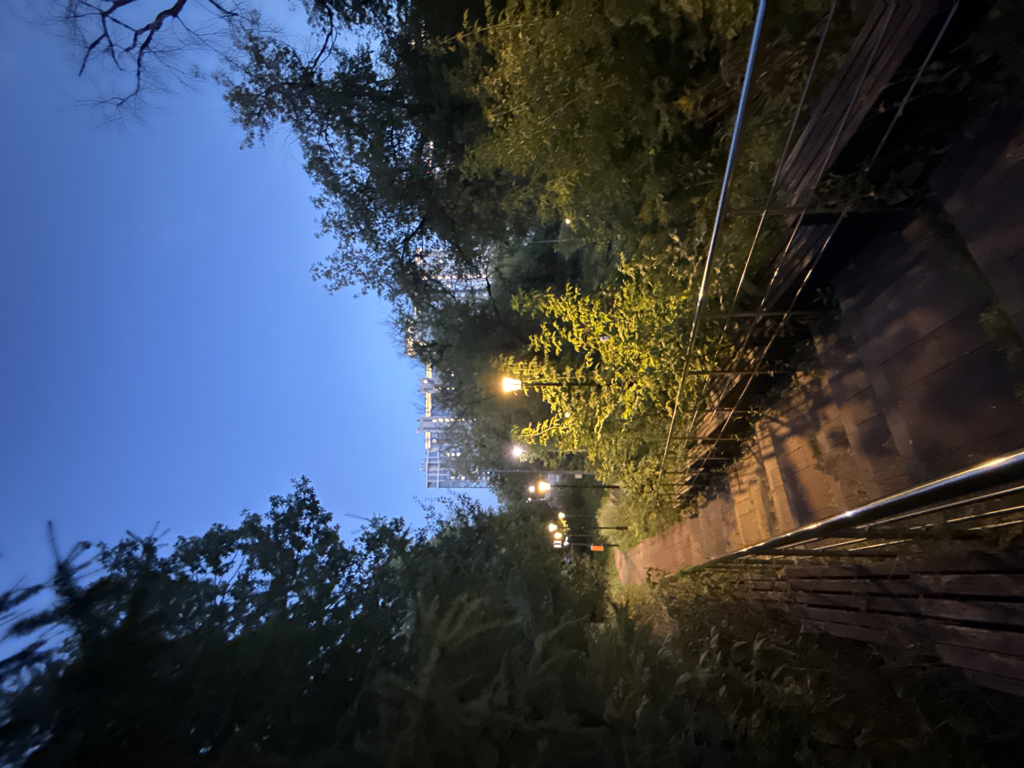 This image depicts a pathway in a park at dusk, with several lampposts illuminating the scene. The pathway is made of wood and stone, and it is lined with metal railings. Trees and foliage surround the path, and in the background, a tall, modern building can be seen. The sky is a deep blue, indicating either early morning or late evening. The overall atmosphere is peaceful and serene, with the lampposts casting a warm glow on the surroundings.