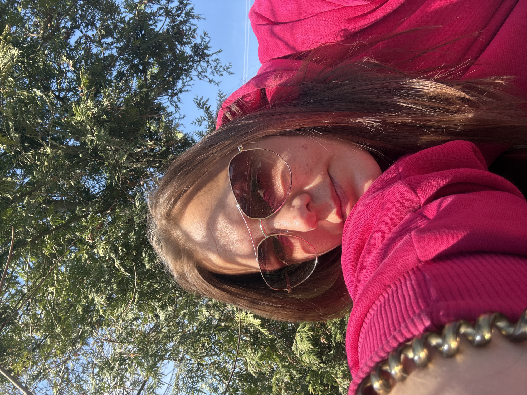 A woman wearing sunglasses and a pink hoodie is seen outdoors, with a tree behind her and a blue sky visible. The presence of specific trees suggests this photograph might have been taken in a park or green space in Dmitrov, Russia.