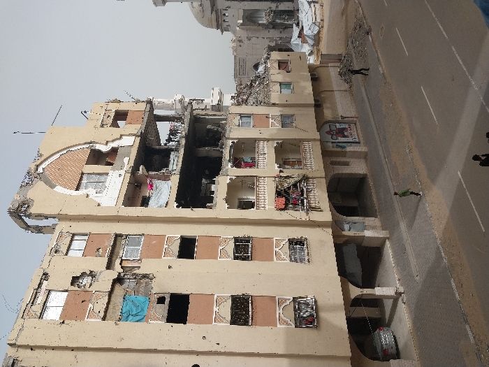 The image captures a severely damaged multi-story building in Khan Yunis, Palestinian Territory, under an overcast sky. The building, primarily tan with brown accents, exhibits extensive structural damage, particularly on its upper floors and right side, where large sections of the facade and roof are missing, exposing interior rooms. Numerous circular impacts, indicative of bullet or shrapnel damage, are visible across the remaining exterior walls. Windows are broken, missing, or covered by materials like a blue tarp on the third floor. Balconies with white railings on lower levels display clothing hanging to dry. The ground floor features arched entryways; one shelters a parked silver sedan, and another has a partially visible storefront poster.
A paved street with white lane markings and a tiled sidewalk run in front of the building. Piles of rubble and debris are present on the sidewalk and extend into the background. To the right of the main building, more heavily damaged structures and the partially collapsed dome of what appears to be a mosque are visible against the skyline, surrounded by further debris. Three individuals are identifiable: one person in a green shirt walks on the foreground sidewalk, another in dark clothing walks further down the sidewalk near a rubble pile, and two more are partially visible at the bottom edge of the frame.