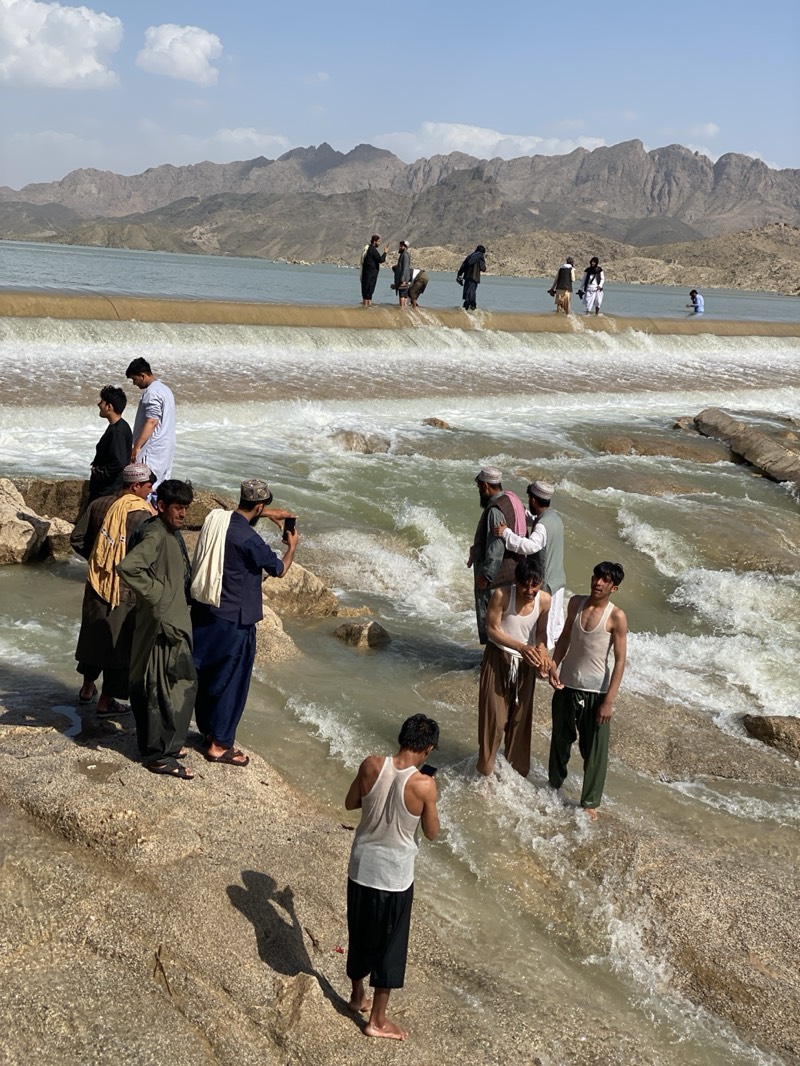 The image depicts a group of men in Kim, Afghanistan. They are gathered on and around a dam or spillway in a river. The men are dressed in traditional clothing, including long robes and head coverings. Some men are taking photos of each other with their phones. In the background, there are mountains and a clear sky. The water is turbulent and flowing rapidly over the dam. Some of the men are standing in the water.