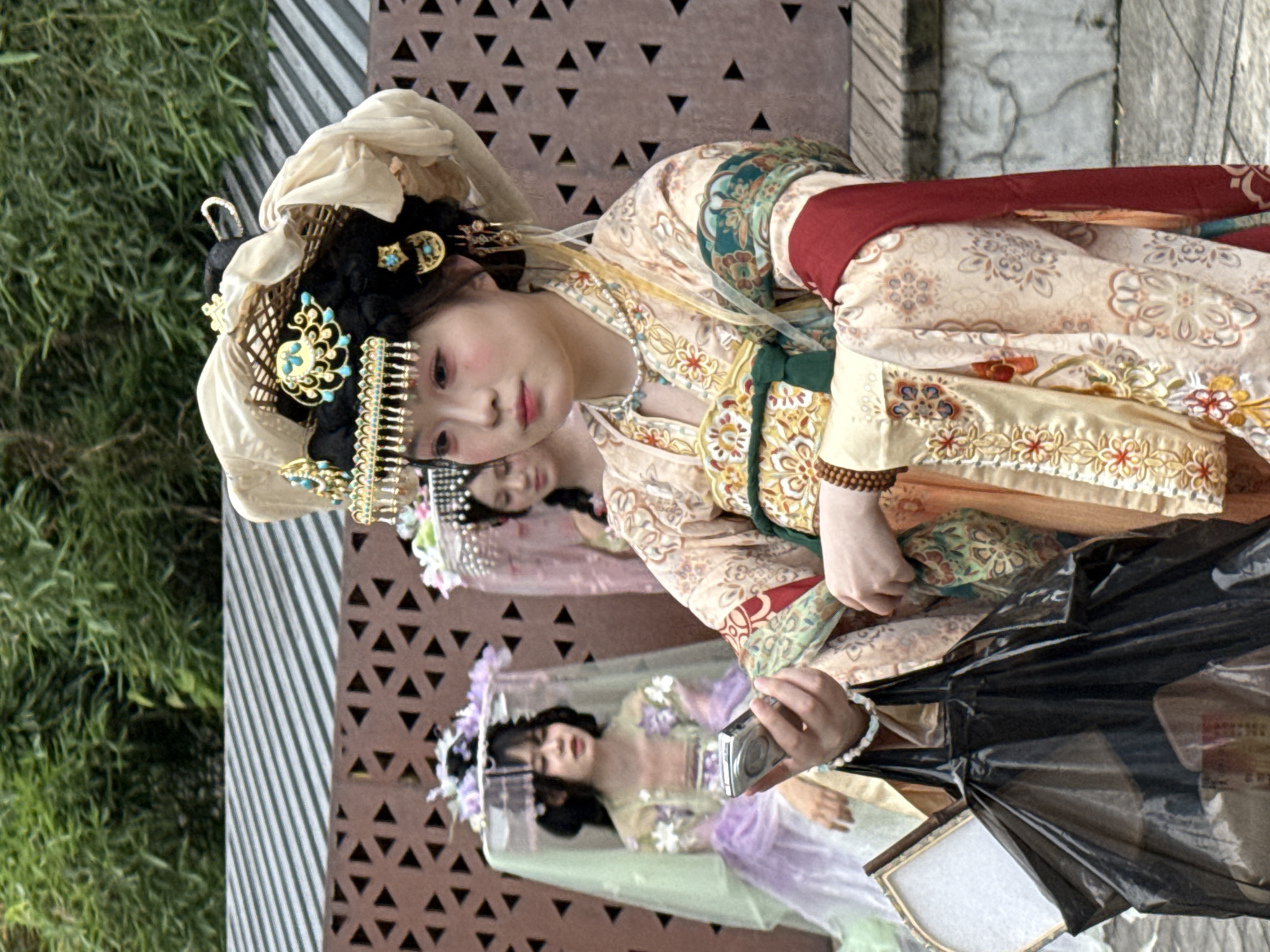 In the Yuzhong District of China, three individuals are captured in a photograph, each adorned in traditional Chinese attire reminiscent of historical Hanfu. The central figure, a young woman, is positioned in the foreground, holding a digital camera in her left hand and a black plastic bag filled with items in her right. Her elaborate headdress features intricate gold and turquoise embellishments, complemented by delicate jewelry around her neck and ears. Her gaze is directed forward, and her makeup is subtle, with a touch of blush on her cheeks. She wears a layered garment with intricate floral embroidery in shades of cream, gold, and red. To her left, another young woman, partially obscured by the central figure, sits facing left, her hands resting in her lap. She wears a light green and purple dress and a veil-like headdress adorned with flowers. Further in the background, a third young woman is visible, also in traditional dress, with a pink veil and floral headdress. The backdrop consists of a textured brown wall with a geometric pattern and a band of horizontal metal slats, all set against a backdrop of lush green foliage.