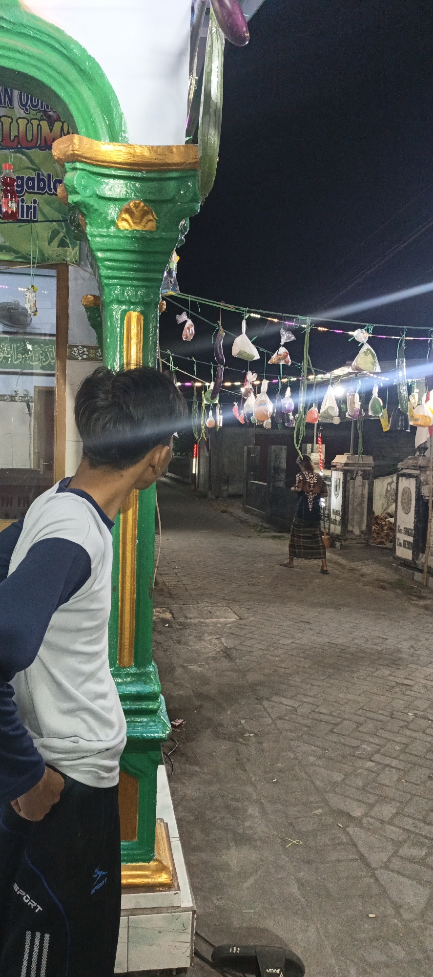 In this nighttime, eye-level outdoor shot, the focus is on a young man seen from the back, turned to his right, observing something in a street. He's wearing a white t-shirt with navy blue sleeves and navy blue track pants with "SPORT" written on them. To his left is a vibrantly painted green and gold pillar, part of what appears to be a building's exterior.
The street itself is paved with cobblestones and illuminated by strings of small, colorful lights overhead, from which various items are hung in plastic bags, including what look like eggplants and produce. A person in traditional clothing, a long patterned skirt and top, walks away from the camera down the street. Buildings line the street, their facades dimly lit, some appearing to be shops or residences. The overall atmosphere is that of a nighttime market or community gathering.