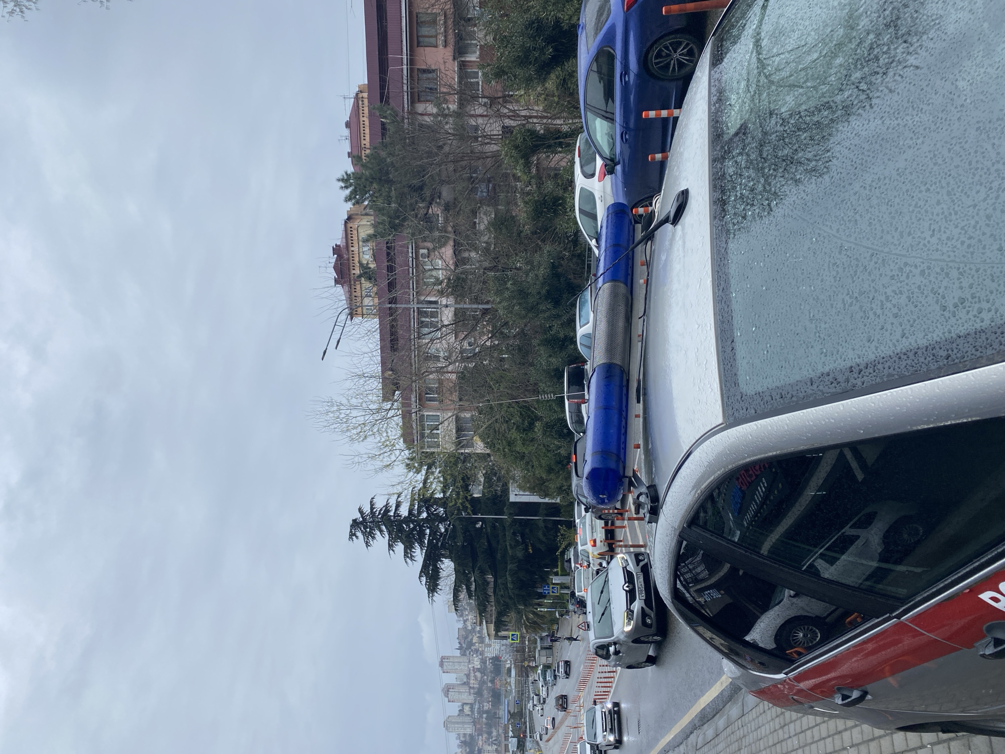 The image captures an urban street scene in Sochi, Russia on a cloudy, possibly rainy day. Cars are parked along the side of the street, with a gray car prominently in the foreground. The windshield and side window of this car are covered in water droplets, partially obstructing the view of the traffic. Behind the row of parked cars, there are residential buildings and trees, giving a partial view of the city skyline. A cleaning vehicle is parked in front of a blue car, with road safety barriers visible in the street. In the distance, city buildings are seen. Traffic is flowing slowly on the street.