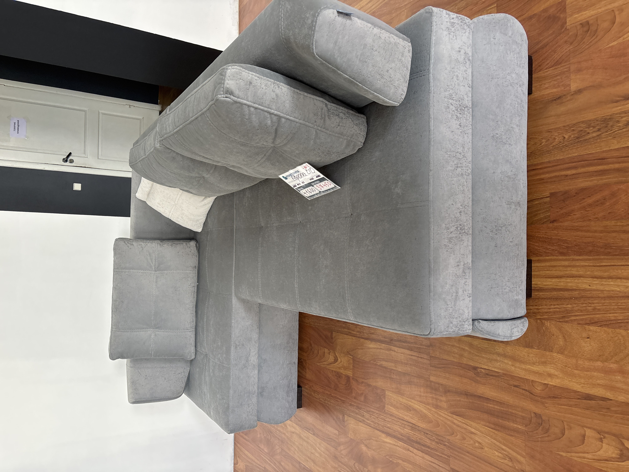 A gray sectional sofa is positioned on a hardwood floor. The sofa has multiple cushions and appears to be in a showroom or domestic setting. A white pillow is placed on the sofa. A price tag is attached to one of the sofa cushions. In the background, there is a white wall and a door with text written on it, and a black wall. The image is taken from a slightly elevated angle.