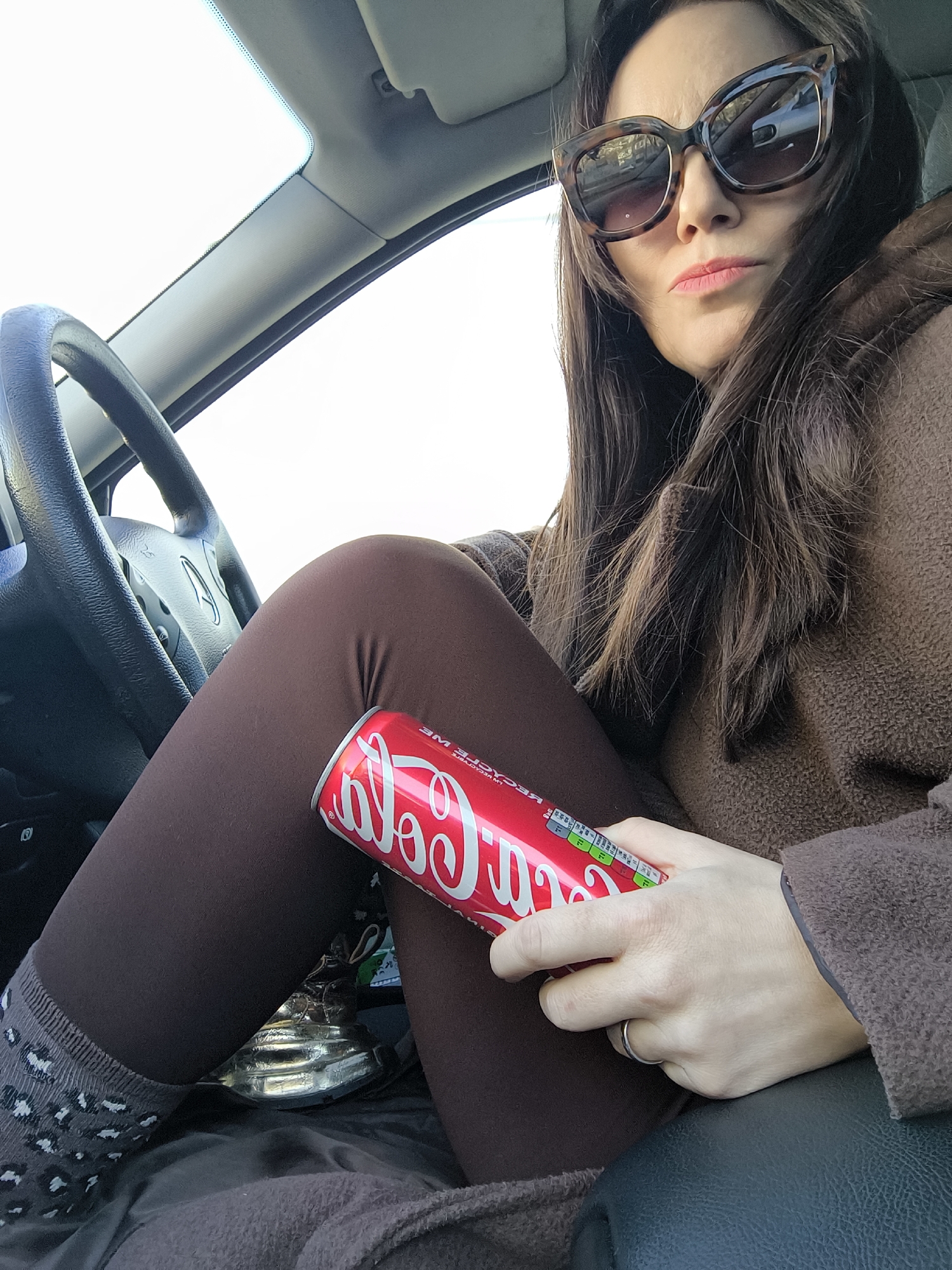 A woman is seated in the front of a car, possibly the driver's or passenger's side, in Riga, Latvia. She is wearing large, tortoise-shell cat-eye sunglasses and a dark brown, textured jacket or coat over brown leggings. Her long, dark brown hair frames her face. Her lips are slightly parted, and she has a neutral to serious expression.
Her right hand holds a red Coca-Cola can, with the iconic white "Coca-Cola" script clearly visible, along with nutritional information and the phrase "RECYCLE ME" (partially visible). She wears a simple silver-colored band on her left ring finger. Her left leg, clad in a brown legging and a grey sock with white, leopard-like spots, is bent. Between her legs, near the floor, a shiny, metallic, possibly decorative object can be seen.
The car's interior features a steering wheel (with an indistinct car logo), part of the dashboard, an A-pillar, and a sun visor. The exterior visible through the windows is extremely bright, suggesting a clear or very bright overcast sky during the daytime. The overall scene suggests a pause during a journey.