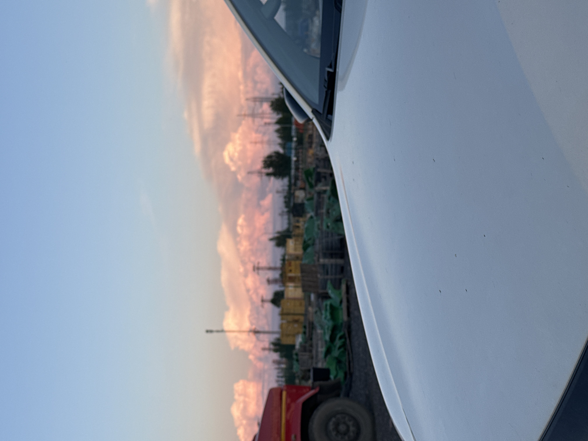 The image depicts a scene in the city of Hora, Ukraine, at dusk. In the foreground, the white hood and windshield of a car are visible. Beyond the car, a red truck is parked on a gravel surface. In the background, there is an industrial area with stacks of green and yellow materials, power lines, and trees. The sky is a gradient of pale blue and pink, with large, fluffy clouds illuminated by the setting sun. The lighting suggests that the photograph was taken during the golden hour. There are no people visible in the image.