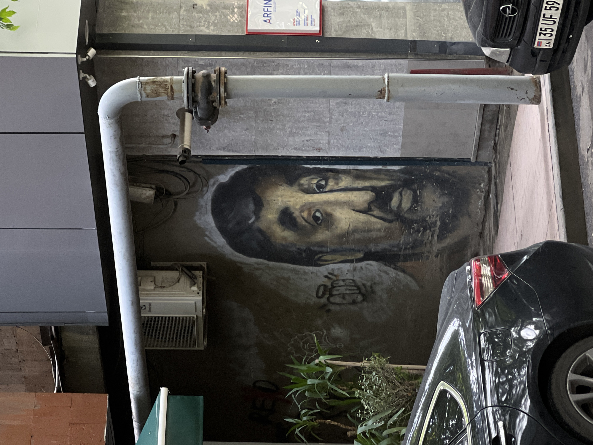 The image depicts an urban scene, likely an alley or side street in Yerevan, Armenia. A large mural of a man's face is painted on a wall. A gray pipe with flanges runs horizontally across the mural. An air conditioning unit is mounted on the wall beside the mural. Below the mural are plants. Two cars are partially visible in the image. The front of a black Opel with a license plate starting with "35 UF 59" is in the upper right corner. The rear of another black car is on the right side of the image. A sign is mounted on the wall above the pipe.