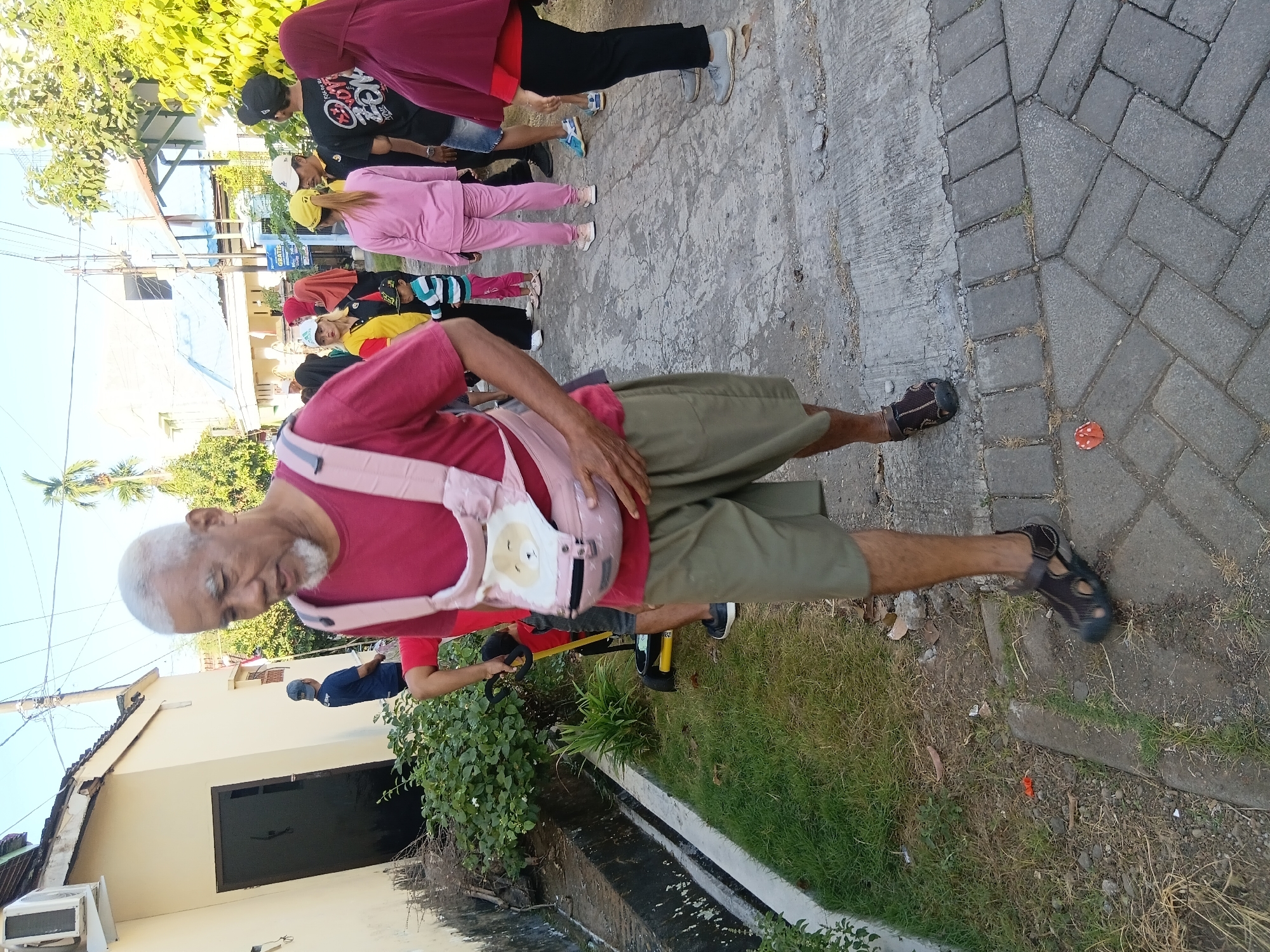 In the city of Tanggul Wetan, Indonesia, an outdoor scene unfolds on a paved walkway adjacent to a grassy area. A man, appearing to be in his late 60s or 70s with white hair and a beard, is walking towards the viewer. He wears a dark red t-shirt and khaki shorts, and a light pink baby carrier is attached to his torso. Several other individuals are visible in the background, some wearing brightly colored clothing, suggesting a gathering or event. The environment includes buildings, trees, and overhead wires, consistent with an urban or suburban setting in Indonesia.