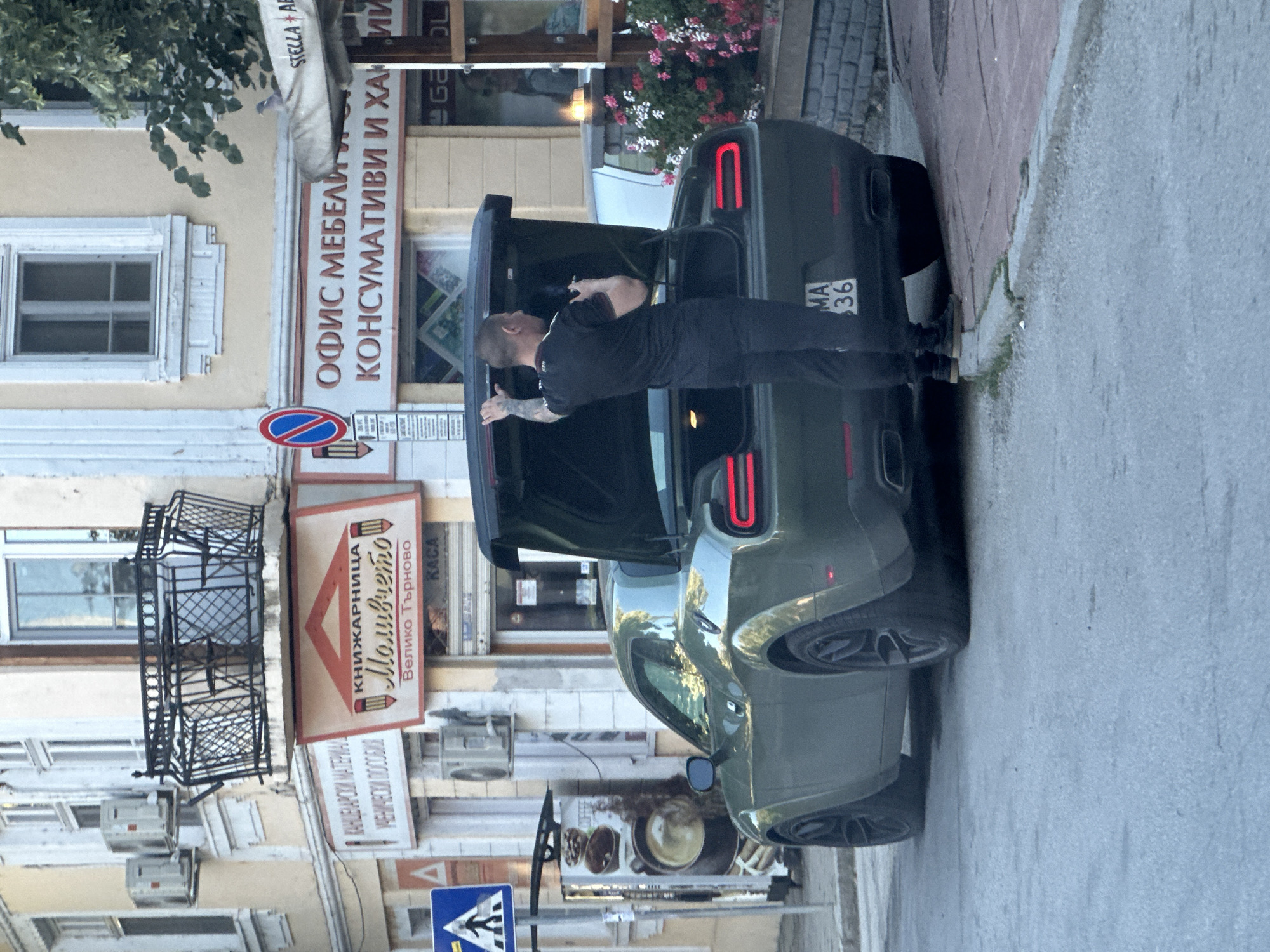 The image captures a street scene in Veliko Tarnovo, Bulgaria. In the foreground, a dark green Dodge Challenger sports car with its trunk open is parked on the right side of the road. A man with tattoos on his left arm, wearing a black t-shirt and black pants, is standing behind the open trunk, holding a black object in his right hand, possibly a phone or a camera. The car's rear taillights are illuminated. To the left of the car, a vending machine with images of coffee is visible, and a pedestrian crossing sign is posted on a pole. Behind the car, the facades of buildings line the street. One building has signage including "КНИЖАРНИЦА Моливчето" (Bookstore Molivcheto) and "Велико Търново" (Veliko Tarnovo), indicating the city. Another sign reads "ОФИС МЕБЕЛИ КОНСУМАТИВИ И ХАНИ" (Office furniture consumables and ...). Air conditioning units are mounted on the building walls. The street pavement is visible in the foreground, with a sidewalk and a brick pathway to the right. Red and white flowers are blooming in planters along the sidewalk.
