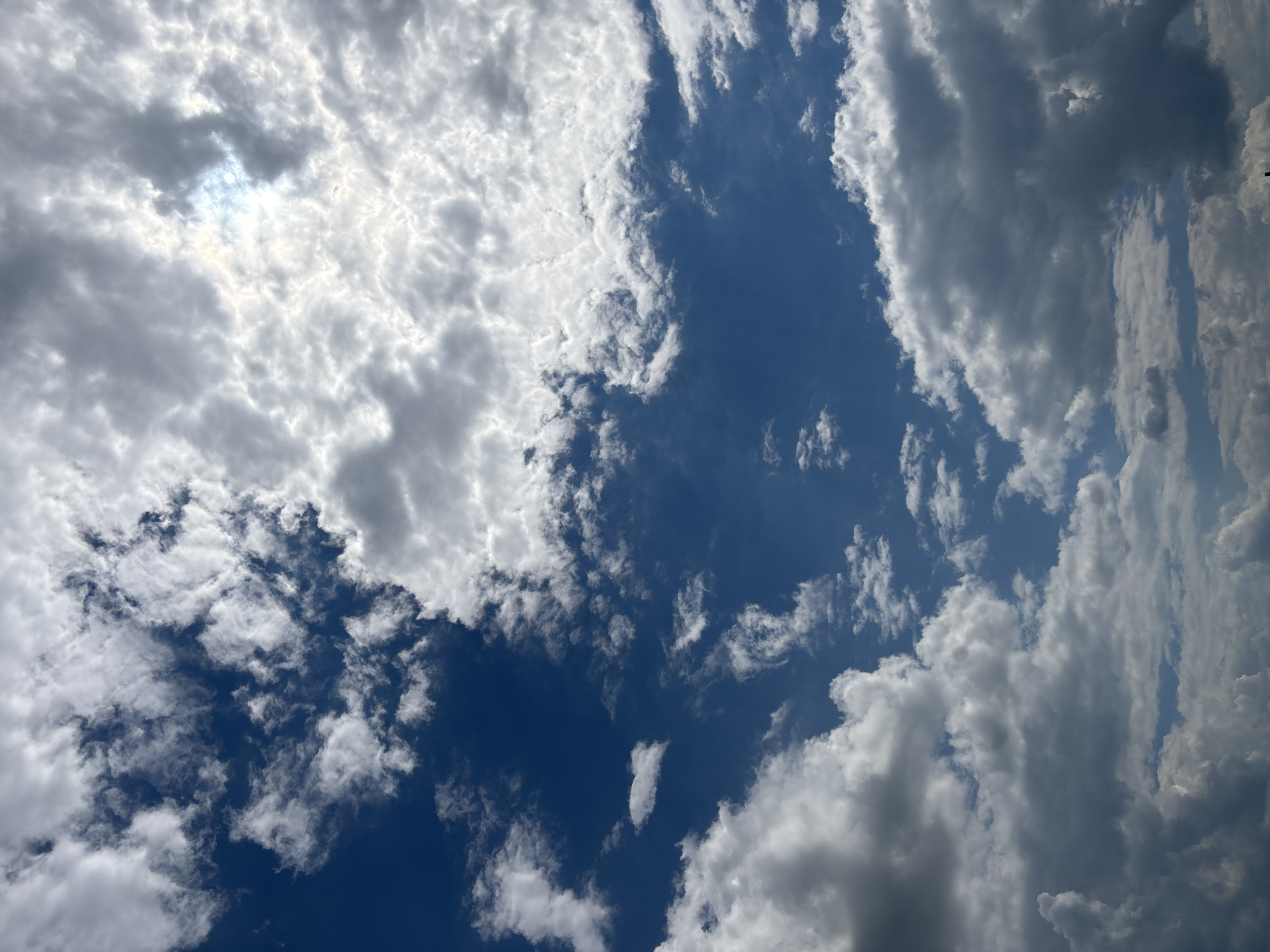 An outdoor, eye-level, full shot captures a view of the sky with varying cloud formations. The sky is a deep blue, with a bright sun peeking through a cluster of white clouds in the upper right quadrant. The clouds range in density and color, from wispy white cirrus clouds to larger, darker cumulus clouds that create shadows. The image appears to be taken from ground level, looking upwards.