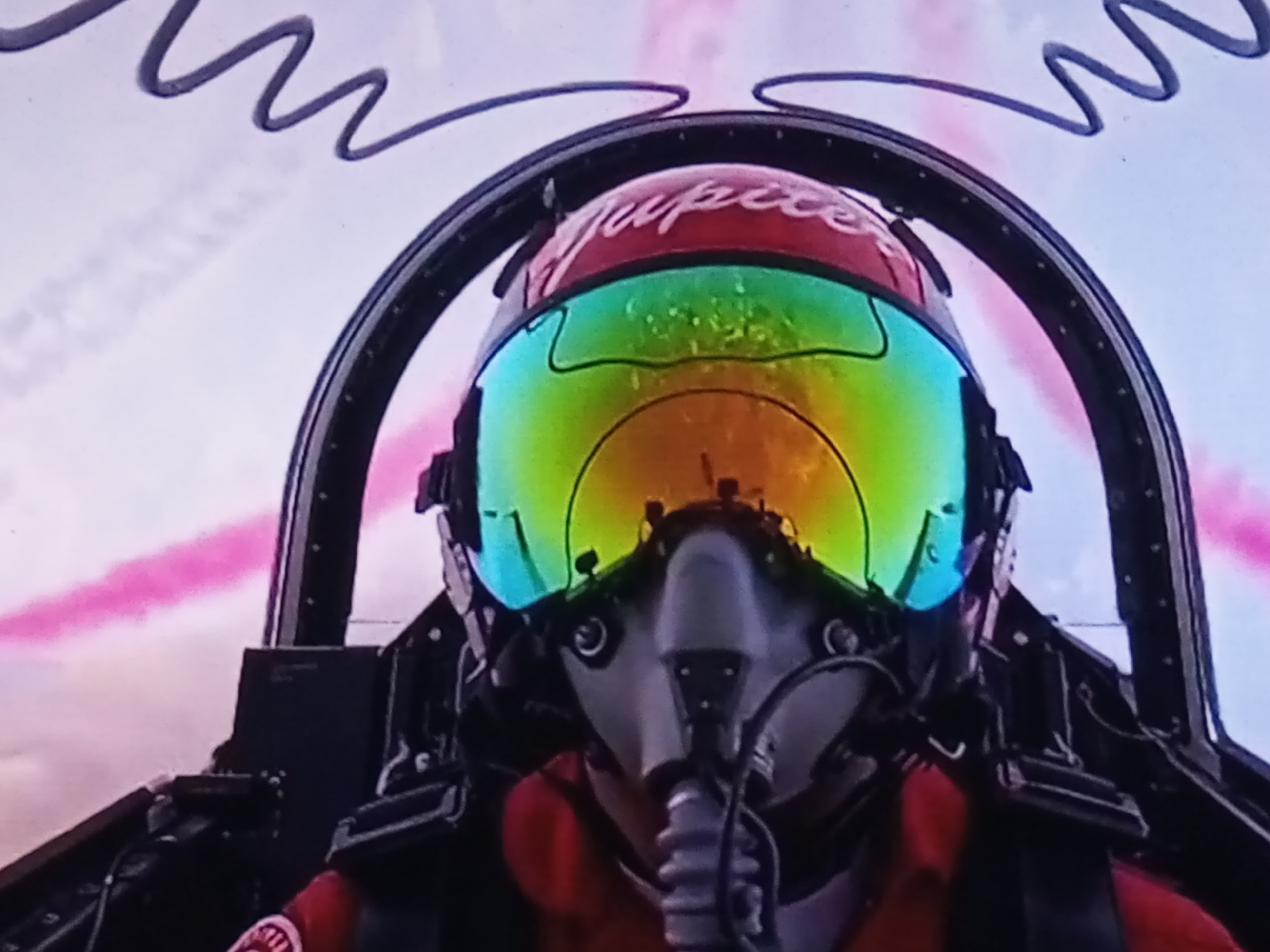 The image shows a close-up, slightly blurry view from inside a jet cockpit, focused on a pilot wearing a helmet and oxygen mask. The helmet is red with the word "Jupiter" written in white script. The visor of the helmet is highly reflective, displaying a rainbow of colors. The pilot is wearing a red flight suit. The cockpit interior is visible, with black panels and some equipment. Outside the cockpit, pink smoke trails are visible against a light-colored sky, suggesting an aerial maneuver or display. There are no discernible geographical landmarks that would indicate a specific location in Banyudono, Indonesia, or any other city.