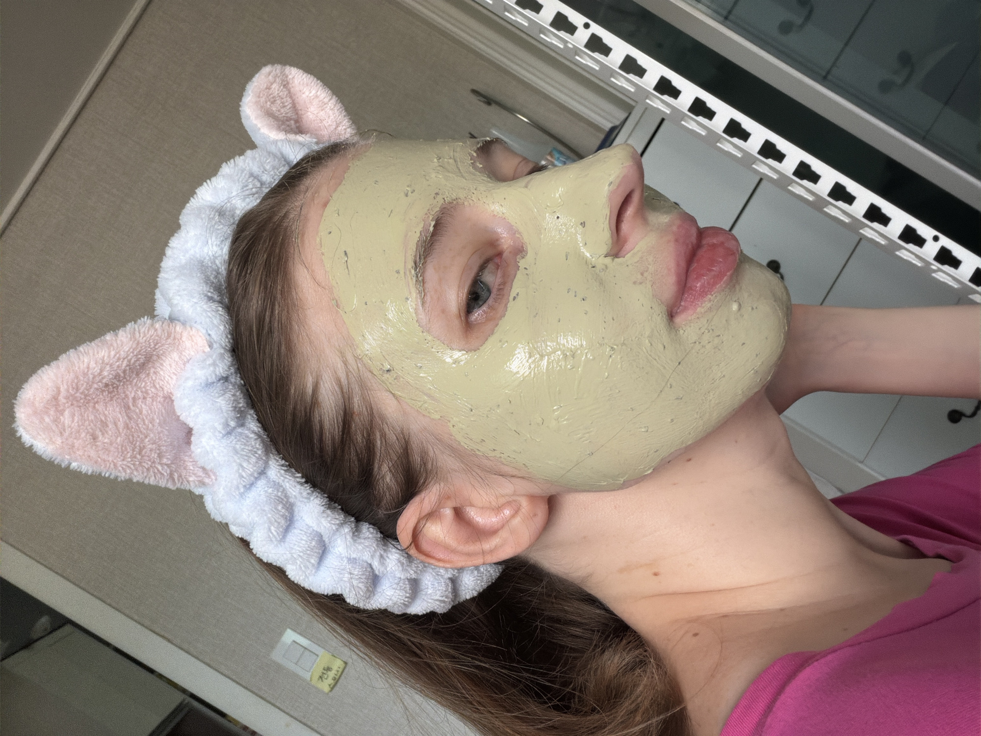A young woman is applying a face mask, she is wearing a fluffy headband with pink bunny ears, and a bright pink t-shirt. The woman has brown hair that is pulled back, and she is looking to her right. The background is an indoor setting with beige walls and white cabinets. There is a vent on the right side of the image.