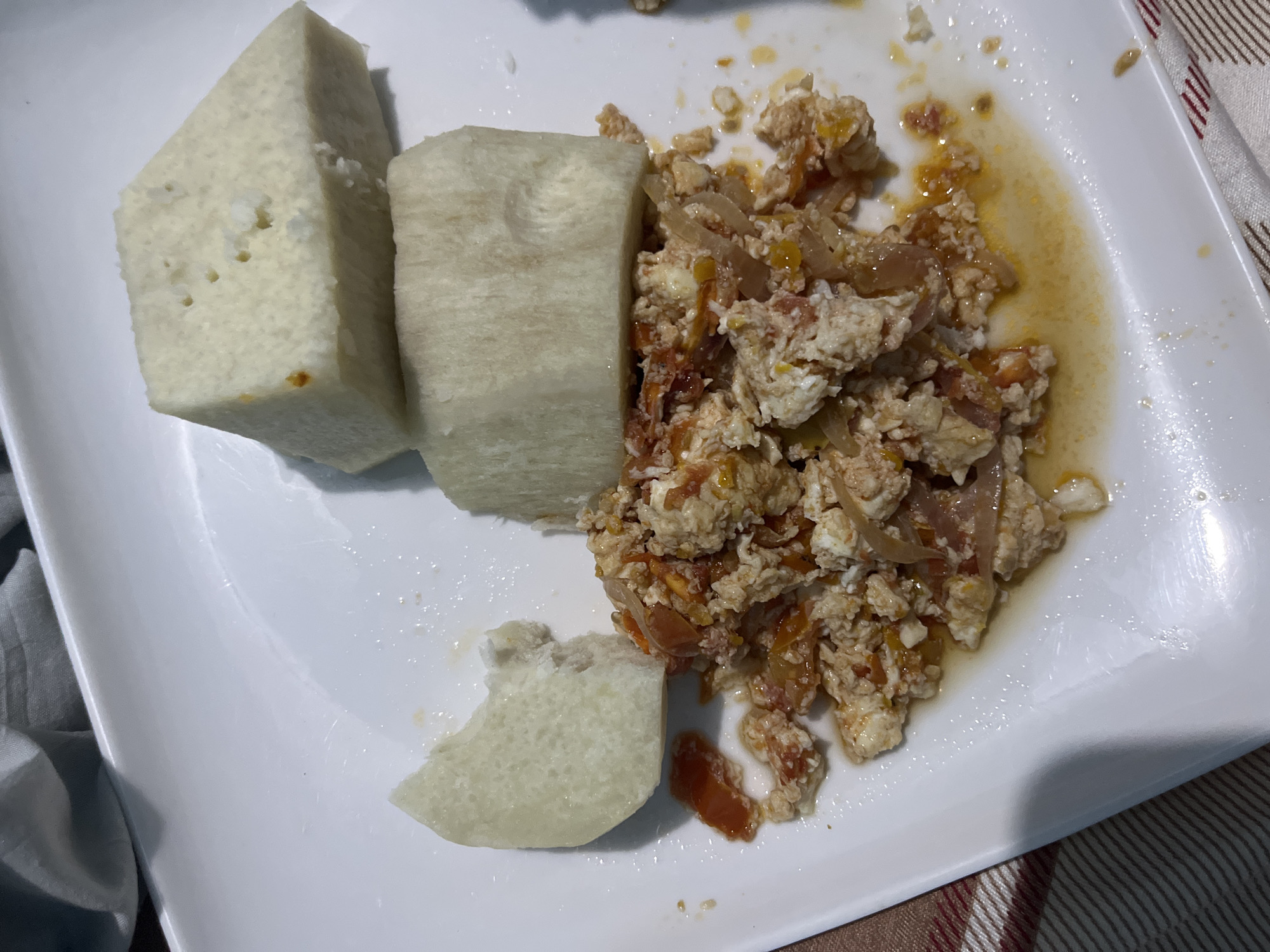 A white rectangular plate holds a portion of yam and scrambled eggs with tomatoes and onions. The yam is cut into three large pieces, with one piece appearing to be a chunk of a larger tuber, and two other pieces being roughly rectangular blocks. The scrambled eggs are a mixture of cooked egg, chopped tomatoes, and sliced onions, with some visible oil and seasoning. The plate rests on a white tablecloth with a striped pattern of red and brown. The image appears to be taken from a top-down perspective, slightly angled. This dish is common in many Nigerian households, including those in Jos, Plateau State.