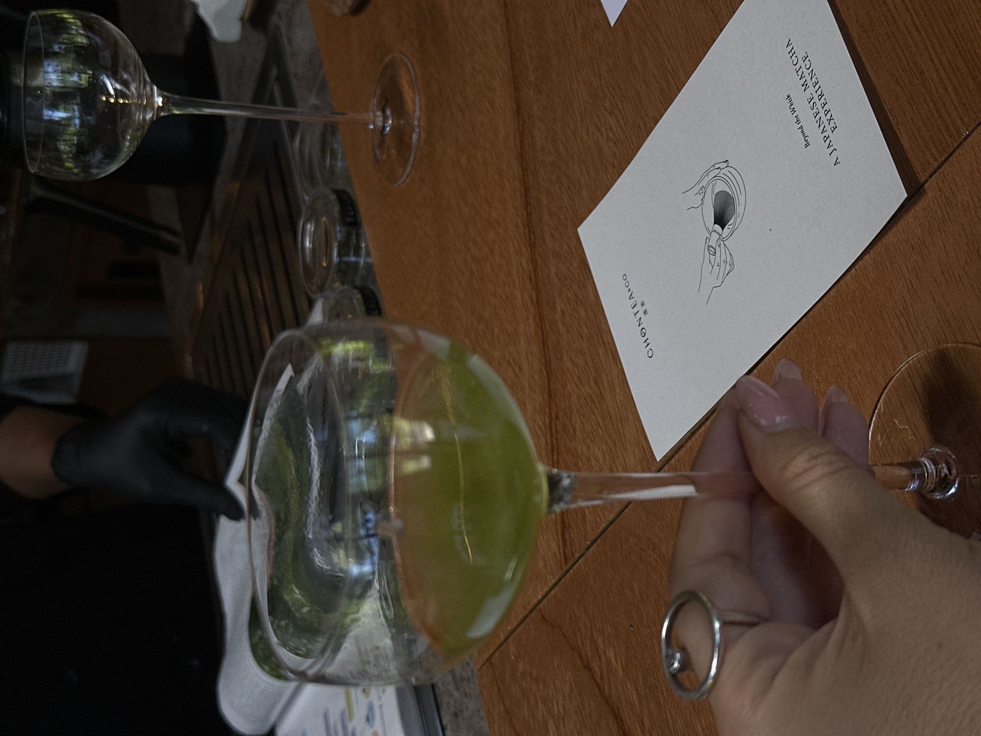 A restaurant scene captured in Ubud, Indonesia, presents a dining table made of wood. On the table is a white card that reads "CHONTEA+CO A JAPANESE MATCHA EXPERIENCE Beyond the Whisk". The photo features a person in a black glove cleaning a wine glass with a white cloth. Another wine glass is positioned above the first, also set on the table. A hand with a ring around one finger is holding the stem of the wine glass.