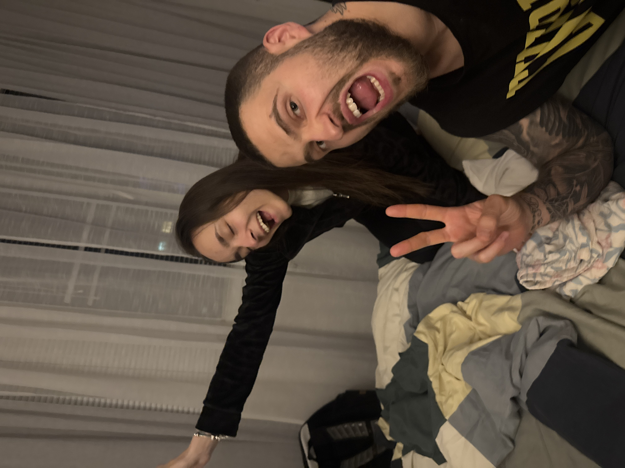 Two individuals are posing for a photograph indoors. A man with a beard, tattoos on his arm, and a black t-shirt is positioned above a woman with long brown hair wearing a long-sleeved black top. Both are smiling widely with their mouths open. The woman is holding up a peace sign with her right hand, and the man is looking towards the camera. They appear to be lying in a bed, with various blankets and a comforter underneath them. In the background, there is a window with blinds. The image does not provide enough information to determine a specific location in Sofia, Bulgaria.