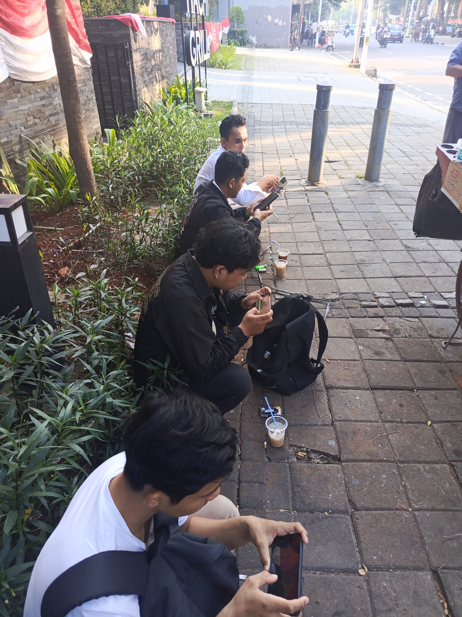 Four young men are sitting on the sidewalk in front of a building, which appears to be a restaurant or cafe, in the city of Central Jakarta, Indonesia. The men are dressed casually, and two of them are holding cell phones. One man in the foreground is holding a cell phone up to take a picture or video. There are several cups of iced coffee on the ground, along with some cigarette packs and butts. In the background, there is a street with traffic, including motorcycles. The building has a brick facade and a large sign with text that is partially visible. There are also some plants and trees in the foreground. The scene appears to be a typical urban outdoor setting.