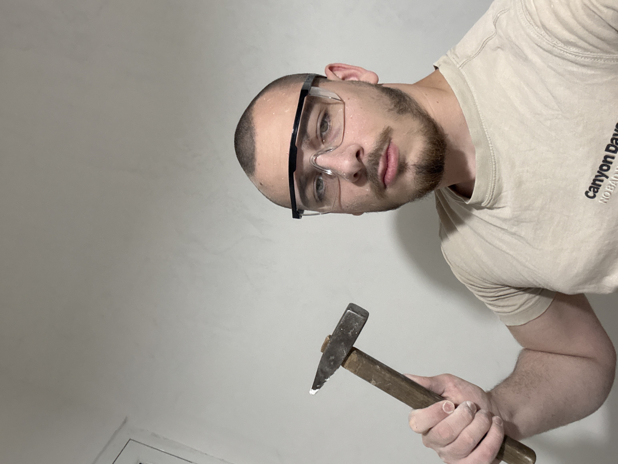 A man is holding a hammer. He is wearing safety glasses and a tan t-shirt with "Canyon Days NO BAD" printed on it. He has a shaved head and a beard. The location is not specified, and therefore unknown.