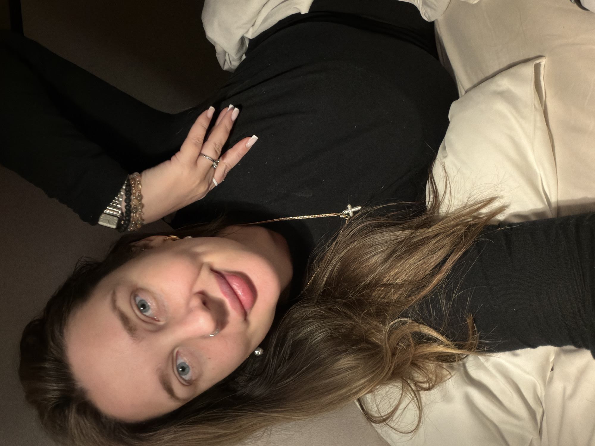 An image, oriented diagonally, features a young person, likely female, lying on a bed in an indoor, dimly lit setting. She has light brown hair, which is spread around her head on a light-colored pillow and bedding. Her blue eyes are open and directed towards the camera, and she has a soft, relaxed smile.
She is wearing a black long-sleeved top. Accessories include a silver-toned ring on her left index finger, multiple beaded bracelets (some dark, some light/gold-toned) on her left wrist, and a watch on the same wrist. A small hoop nose ring is visible on her right nostril, and she wears subtle stud earrings, one appearing pearl-like. A gold-toned necklace with a small cross pendant hangs around her neck. The background is indistinct and dark, consistent with low light conditions, suggesting evening or night. No specific location cues are visible within the image, but the provided context places the scene in Moscow, Russia. There is no visible text in the image.