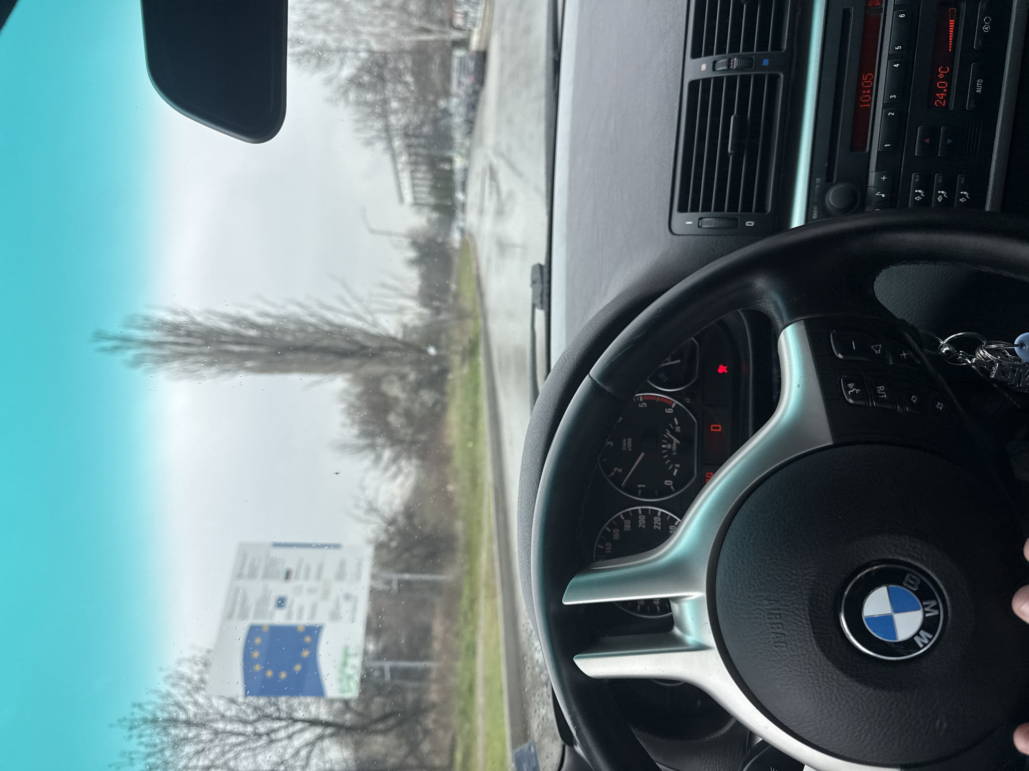 The image depicts the interior of a BMW car, specifically the dashboard, steering wheel, and a view through the windshield. The car's dashboard features a climate control panel displaying "24.0 °C" and a radio showing "10:05". The steering wheel prominently features the BMW logo. The view through the windshield shows a road and signage, with a notable sign displaying the European Union flag and other information, potentially indicating a border crossing or a location with EU-related information. The environment outside appears somewhat overcast or rainy. The location is identified as Sofia, Bulgaria.