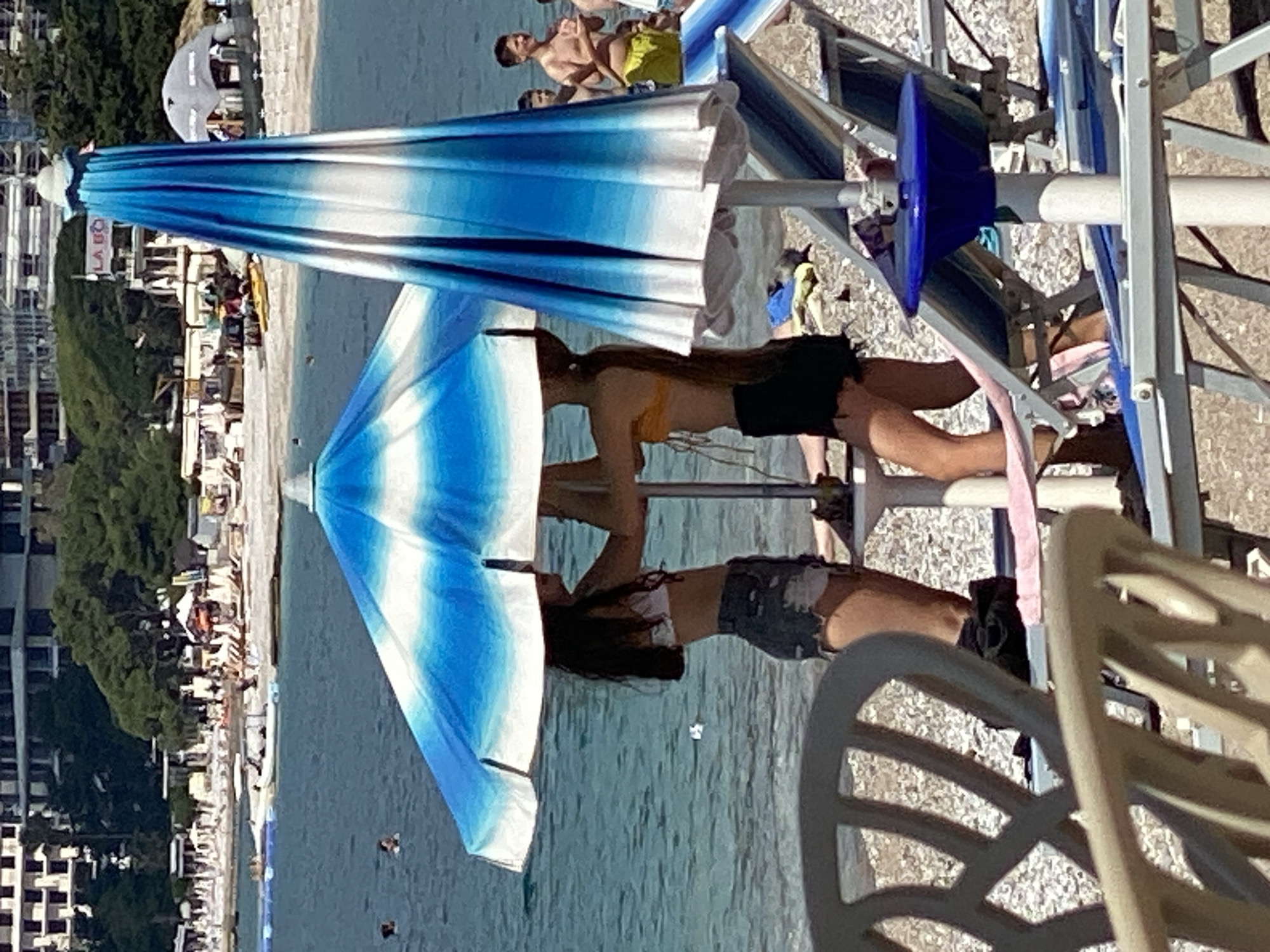 The image shows a beach scene in Budva, Montenegro. In the background, there are buildings and trees indicative of an urban area adjacent to the coastline. Along the beach, numerous sun umbrellas are visible, with blue and white stripes. Two women are on beach chairs under an umbrella. Other people appear in the background. The beach is lined with sunbeds and chairs. The water looks calm and clear. A white plastic chair is in the foreground.
