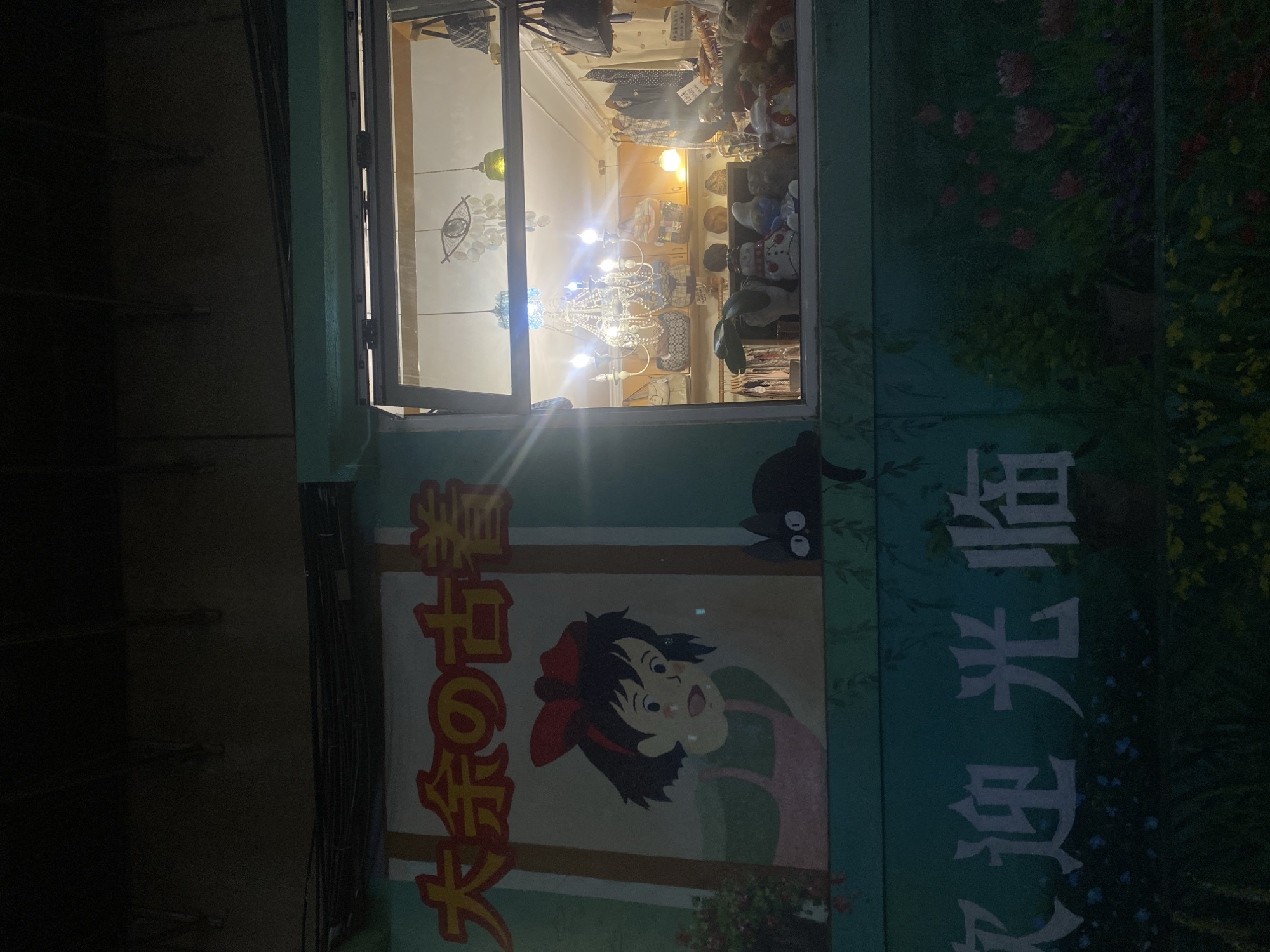 This is a nighttime exterior shot of a shop in Kunming, China. The shop appears to be a vintage or second-hand clothing store, as indicated by the Japanese text on the signage which translates to "Grandma's Second-Hand Clothing" and a mural of an anime character, possibly Kiki from Kiki's Delivery Service, alongside a black cat. The shop window displays a chandelier and various items for sale, including clothing, bags, and stuffed animals. The lower part of the building features a mural of flowers and foliage. The overall ambiance is somewhat whimsical and nostalgic, illuminated by the shop's interior lighting and possibly streetlights, though the surrounding environment is dark.