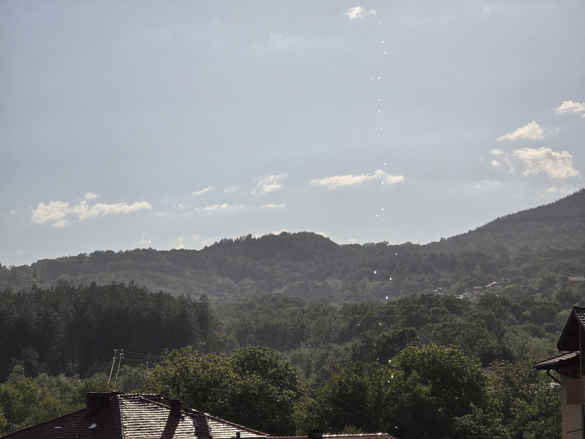 The image shows a landscape in Troyan, Bulgaria, featuring a view from an elevated position towards a wooded hillside with scattered houses. The foreground is dominated by rooftops of buildings, with trees partially obscuring the view. In the middle ground, a densely forested hillside rises towards the sky. In the background, the sky is overcast with clouds. A series of vertically aligned bright spots are visible in the sky. These spots are most likely lens flares or reflections. The overall lighting suggests a daytime scene.
