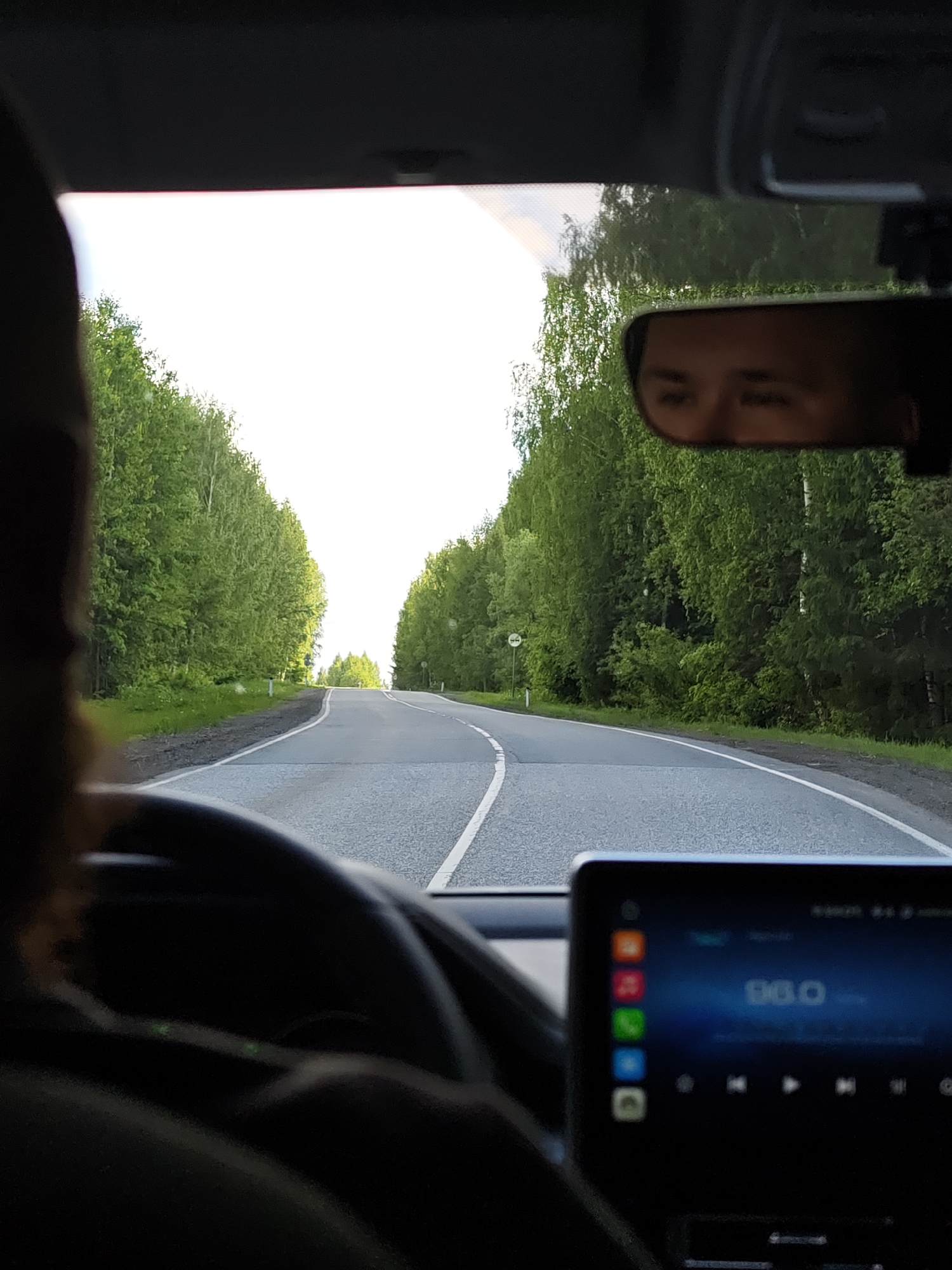The image shows a view from inside a car driving on a paved road lined with green trees, likely in a rural or semi-rural setting. The road curves slightly ahead. A white dividing line runs along the center of the road.
The person is driving in the car, as suggested by the presence of a steering wheel and the reflection of their face in the rearview mirror.
The car's infotainment screen is visible, displaying what appears to be a radio station (96.0).
A sign is visible along the side of the road, though its details are not clear enough to read.
I am unable to identify the location in the city of Oralki, Russia as there are no specific landmarks in the image to do so.