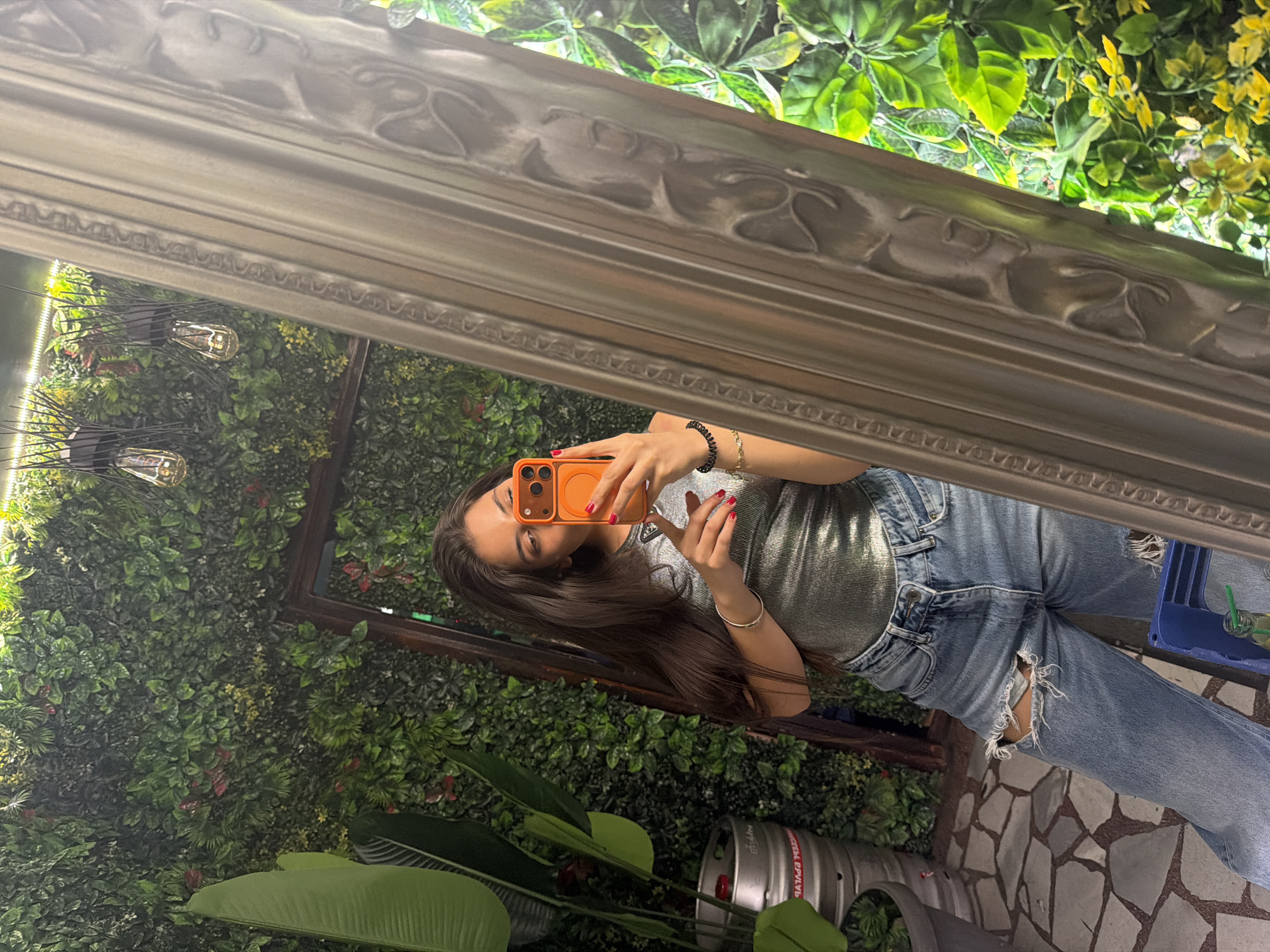 A young woman is taking a mirror selfie in a location that appears to be a bar or restaurant, based on the presence of a beer keg in the lower part of the frame. The interior space is adorned with abundant greenery, including a wall covered in various types of foliage and some large, broad-leafed plants in the foreground. Decorative lighting fixtures can also be seen on the green wall.
The woman is holding an orange-cased smartphone, seemingly capturing her reflection in an ornate-framed mirror. She is wearing a shimmering silver top and light blue denim jeans that are ripped at the knees. Her hair is long and dark.
The location is potentially in Rudartsi, Bulgaria, based on the request, though this cannot be verified from the image alone. The floor is tiled with irregular stone-like shapes. There is a blue crate or tray visible to the right, containing what appears to be some drink-related items.