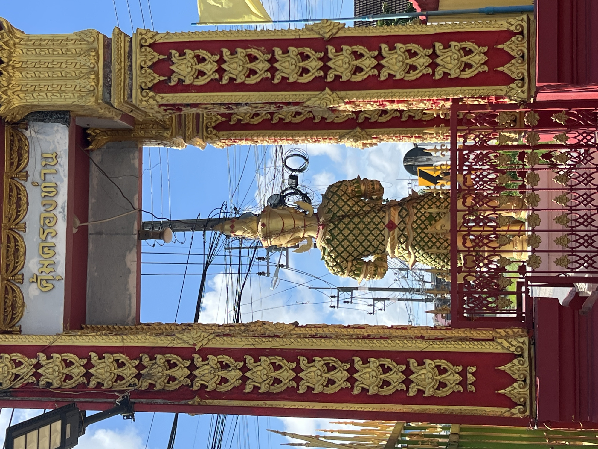 The image shows a golden statue of a mythical guardian figure positioned in front of a decorative red and gold archway. The statue, viewed from the back, is adorned with a green and gold patterned garment and a tall headdress. The archway features intricate golden ornamentation against a red background. In the background, a blue sky with scattered clouds is visible, along with numerous power lines and utility poles. A yellow flag and part of a building are also present on the right side of the frame. The scene appears to be outdoors in a public area within the city of Thep Krasatti, Thailand.