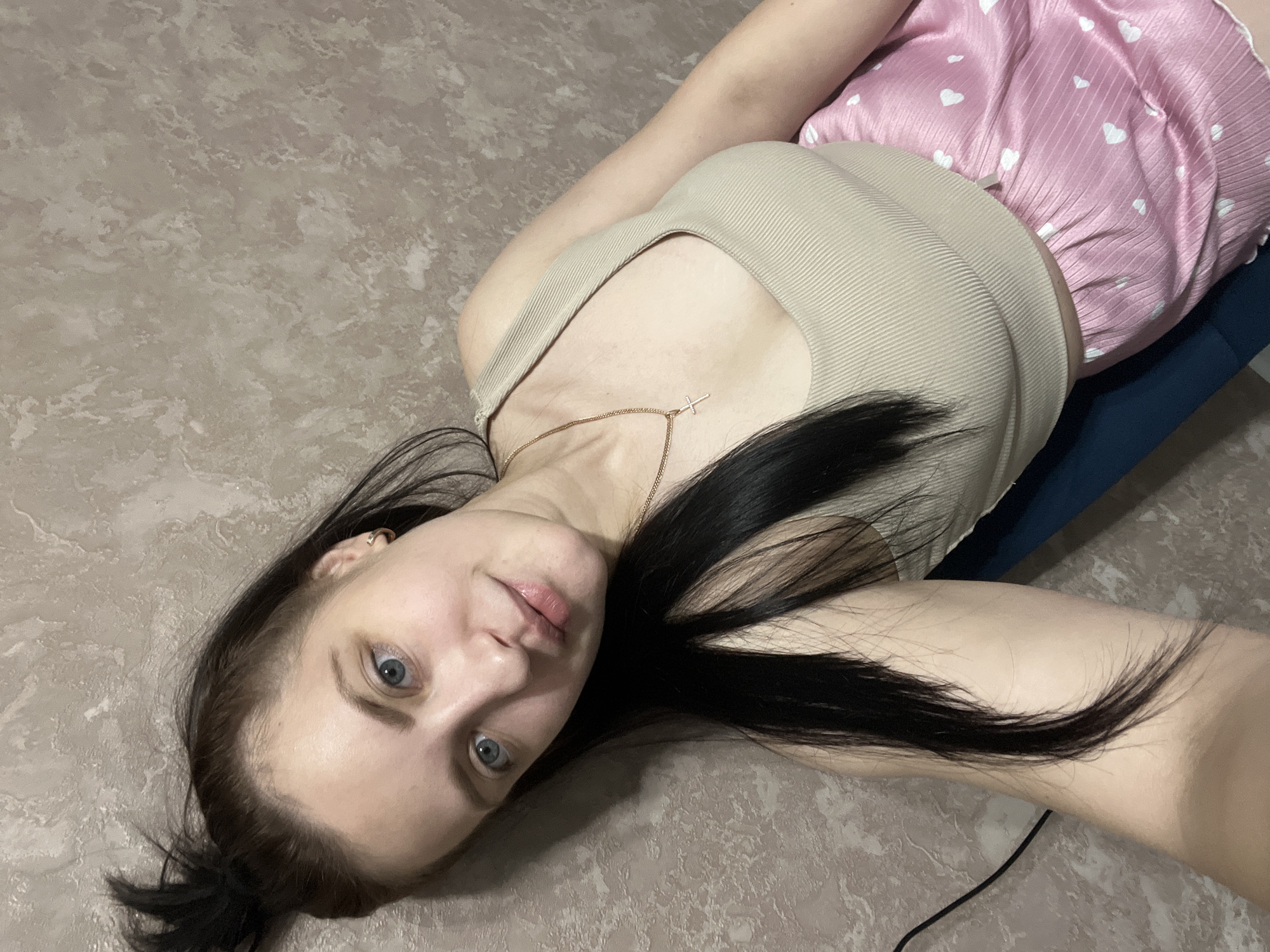 A young woman with long, dark hair and blue eyes is lying on a patterned floor. She wears a beige tank top and pink shorts with white heart patterns. A gold necklace with a cross pendant is visible on her chest. A dark blue object appears behind her right shoulder. The scene is indoors. The city of Марковский, Russia, is not discernible from this image.
