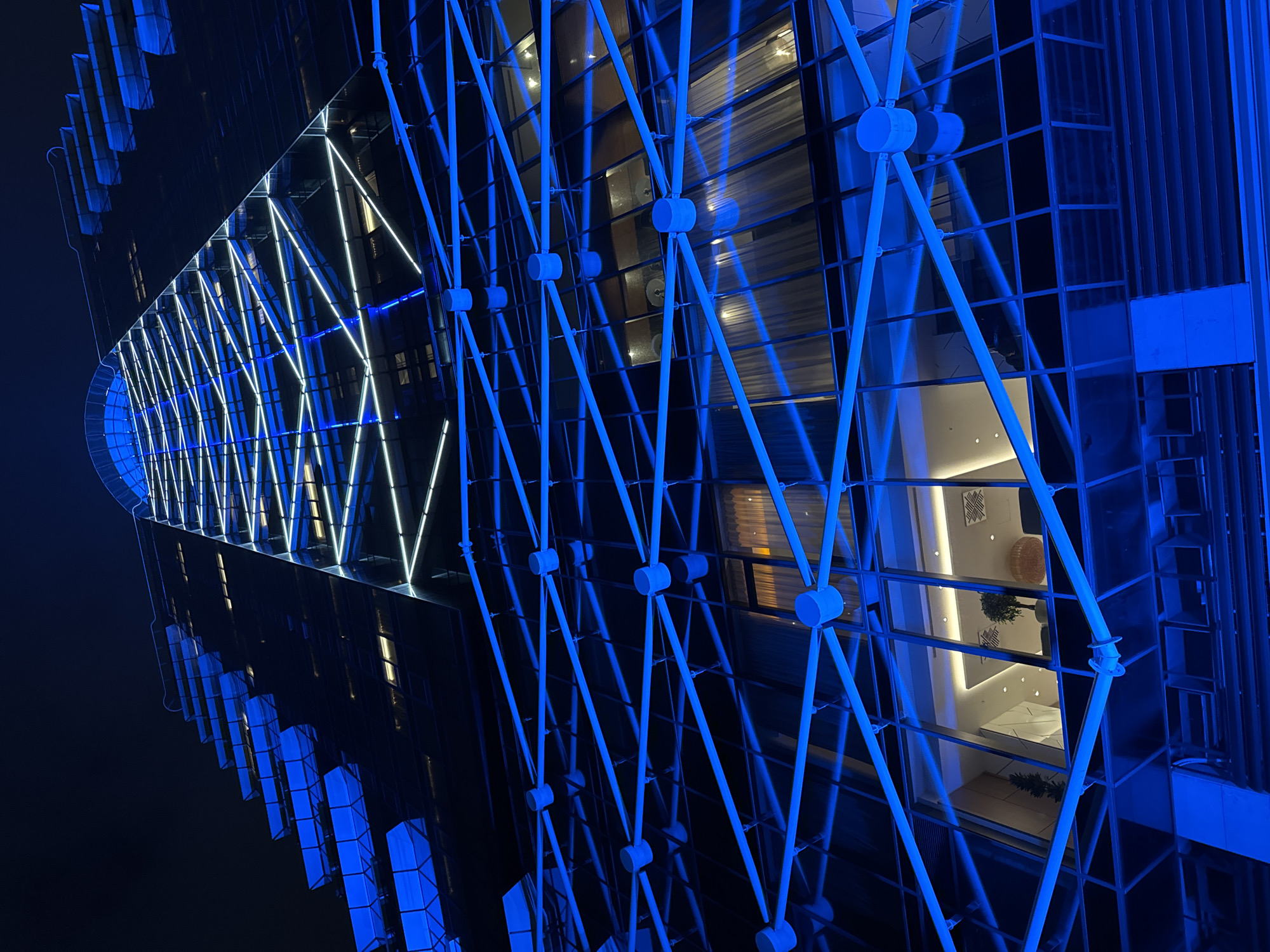 The image displays the exterior of a modern building at night, located in Sofia, Bulgaria. The primary subject is the building's striking facade, characterized by large glass panels and an intricate blue metal framework that forms a geometric, almost diagonal, lattice pattern across the surface.
A notable feature is a long, diagonal section on the upper part of the building, illuminated by bright white LED strip lights arranged in a continuous series of triangular shapes, creating a dynamic visual accent. The deep blue hue of the metal structure contrasts with the stark white of the LEDs and the dark night sky.
Through the glass panels, reflections of interior lighting and other parts of the building are visible. In several sections, glimpses into internal spaces reveal warm, softly lit rooms with minimalist decor, including a potted plant and a woven basket. The overall impression is one of sophisticated, contemporary architecture designed for an urban environment. There is no discernible human activity or visible text in the image.