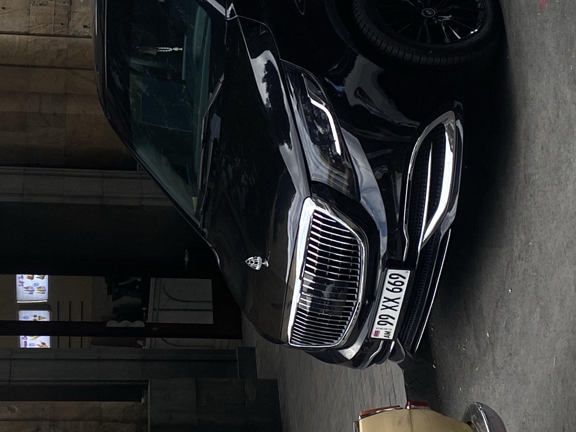 The image displays the front-left portion of a black luxury vehicle, strongly resembling a Mercedes-Benz Maybach, parked in what appears to be an urban setting. The car's prominent feature is its large, chrome grille with vertical slats, reflecting light and surroundings. Above the grille, a distinct Maybach emblem is visible on the hood. The vehicle's sleek, angular LED headlights are partially captured on the driver's side.
An Armenian license plate, "99 XX 669," is clearly affixed below the grille, featuring the country code "AM." Part of a black multi-spoke wheel is visible on the car's right side. In the lower foreground, a section of an older, lighter-colored car, possibly beige or yellow, with a chrome bumper, is partially visible.
The background includes a building with a stone-block facade. Through a window or opening in the background, several illuminated rectangular signs with various graphic designs are visible, suggesting an indoor commercial space. The scene appears to be set during the daytime, possibly in a shadowed area or on an overcast day, judging by the ambient light and reflections on the car's glossy finish. No people or specific activities are depicted, portraying a static street or parking scene in Yerevan, Armenia.