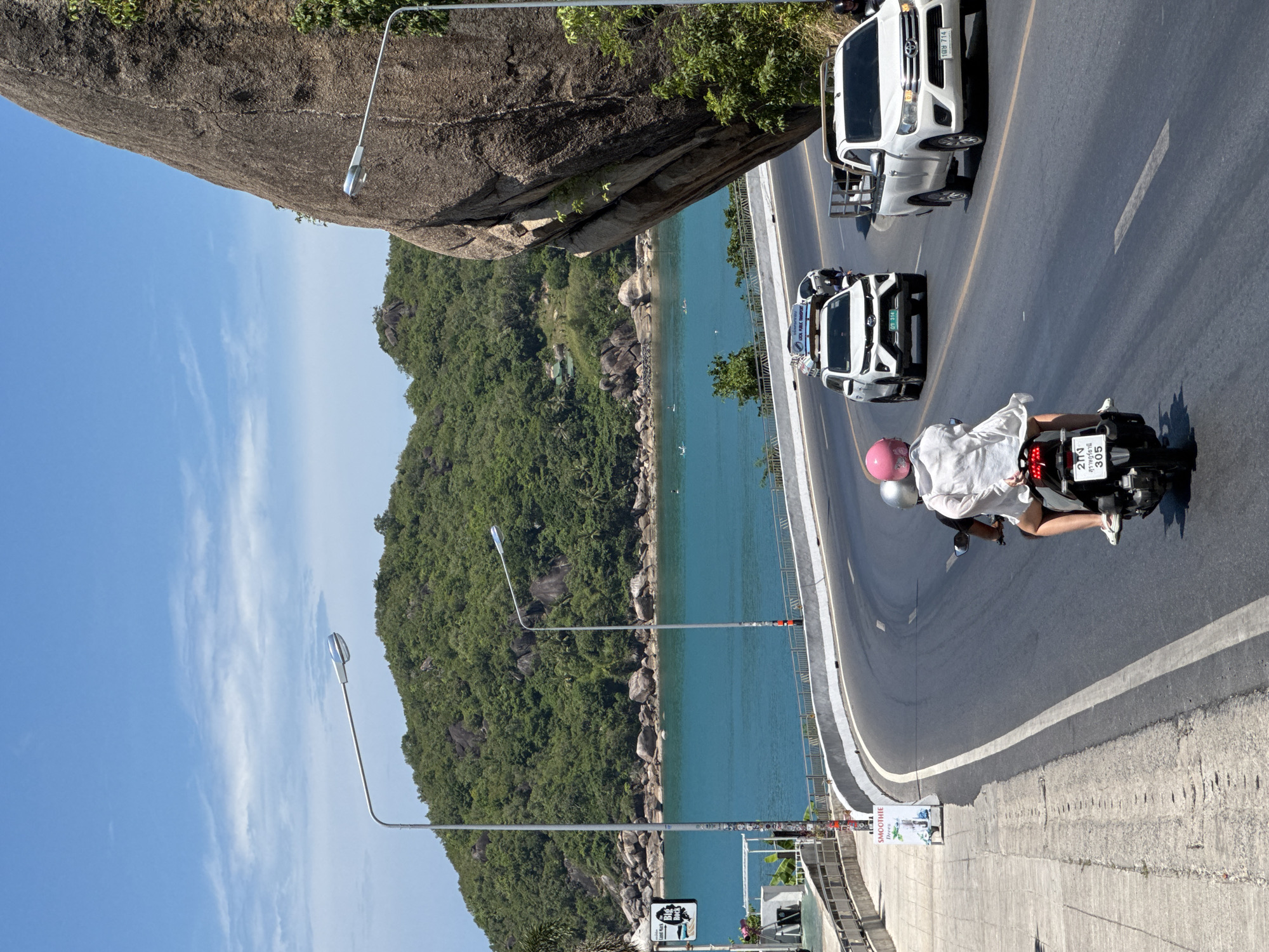 A high-angle, full-shot photograph captures a bright, sunny day on a winding coastal road in Ko Samui District, Thailand. The turquoise ocean is visible to the left, bordered by a lush, green, tree-covered hillside dotted with rocks. A massive, weathered grey boulder dominates the right side of the frame, leaning over the road. Two people are riding a black motorcycle in the foreground, with the passenger wearing a white shirt and a pink helmet. The motorcycle has a Thai license plate. Ahead of them, a white SUV is traveling in the same direction, also with a Thai license plate. Another white pickup truck is further ahead on the right side of the road. A white street lamp with a curved pole is visible on the left side of the road, and another similar street lamp is positioned near the large boulder on the right. The road has a white center line. A paved sidewalk runs alongside the left edge of the road, with a sign that reads "SMOOTHIE DRESS." The sky is a clear blue with sparse white clouds.