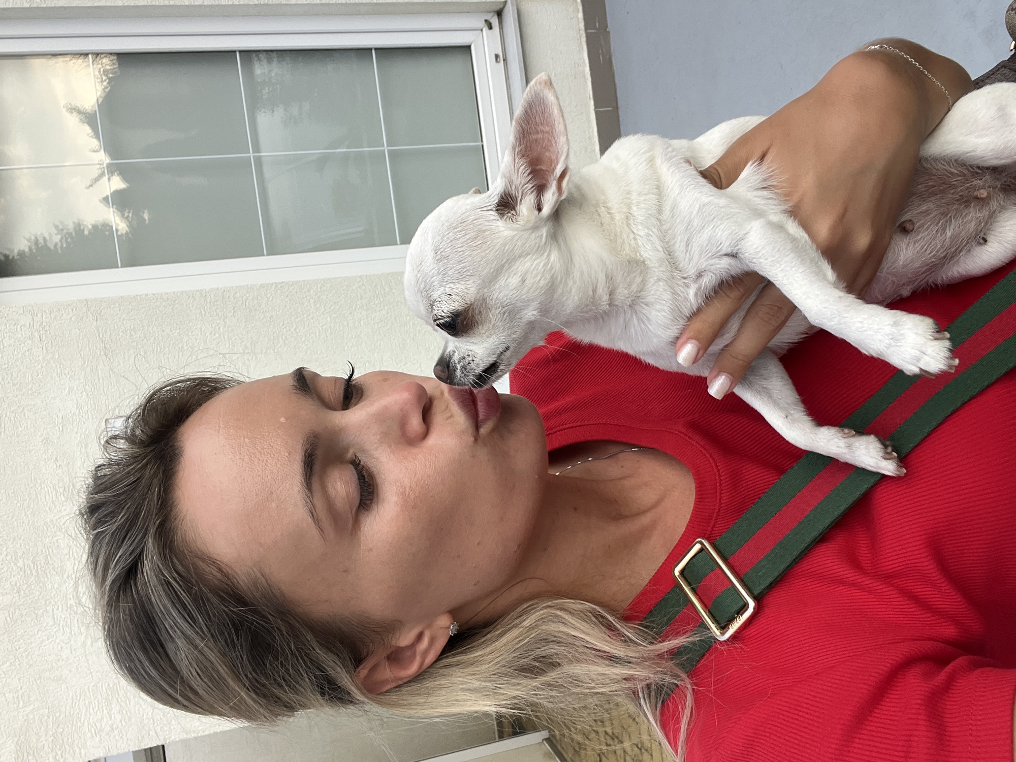 A woman with blonde hair pulled back is holding a small white dog and leaning in to kiss it. She is wearing a red shirt with a green and red striped strap over her shoulder. The background shows a textured white wall and a window with panes. The ground is a grey surface. The scene appears to be outdoors.