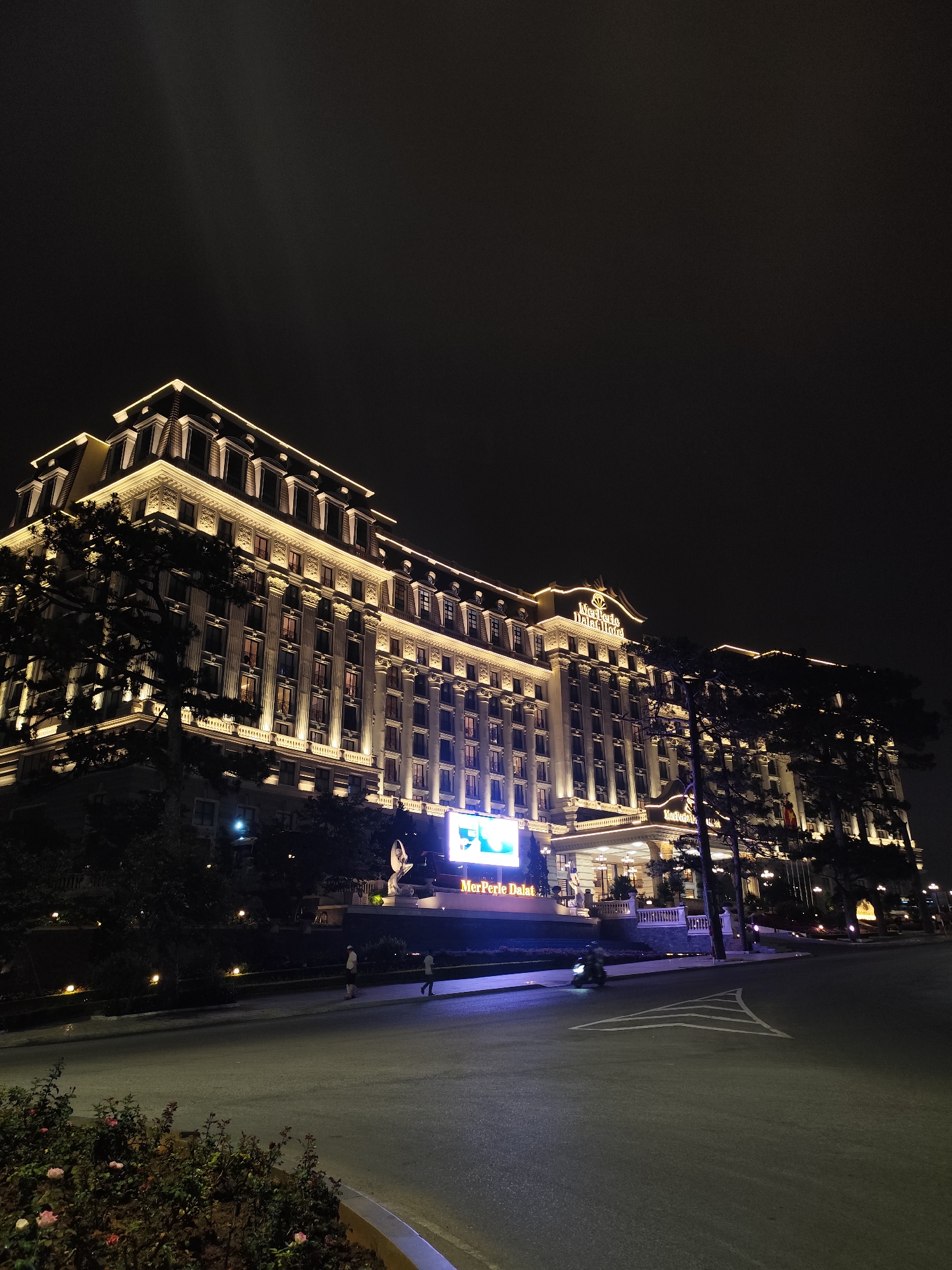 The image captures the illuminated facade of the MerPerle Dalat Hotel in Da Lat, Vietnam, at night. The hotel is a large, multi-story building with a classical architectural style, emphasized by ornate detailing and warm lighting outlining its edges and columns. A bright LED screen displays the hotel's name, "MerPerle Dalat," above the entrance. Below the screen, a couple of people are walking along the pavement. Further into the scene, a motorcycle is on the road in front of the hotel. The foreground shows a section of the road and a garden bed with some low-lying plants and flowers. The sky above is dark and indistinct, typical of nighttime.