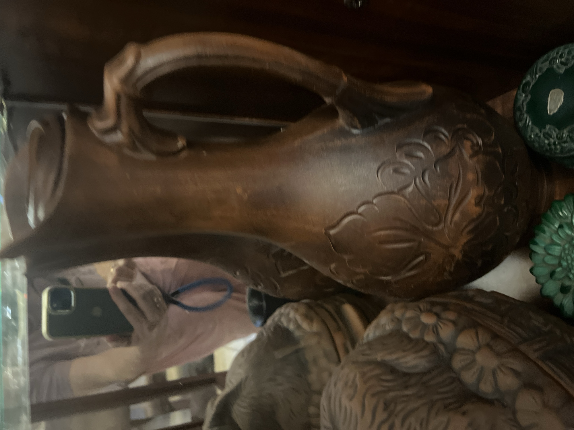 The image shows a dark brown ceramic jug with an ornate handle and floral patterns. The jug is placed on a shelf, partially obscuring other objects. To the left of the jug are other ceramic pieces with textured surfaces, possibly decorative plates or bowls, one of which has a prominent floral design. In the background, a mirror reflects a person holding a smartphone, and a portion of a light-colored garment. A dark wooden surface is visible to the right of the jug, and a small dark object, possibly a cup, is seen behind the jug. Green decorative items, possibly glass, are visible at the bottom of the frame. The scene appears to be an indoor display of decorative items. The location of Vladimirovka, Russia, is not discernible from the image itself.