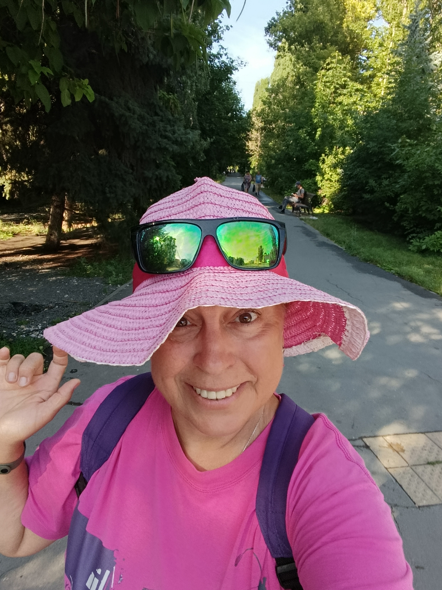 A woman wearing a pink hat and sunglasses takes a selfie in a park. She is smiling and her left hand is raised. She is wearing a pink t-shirt and a purple backpack. The background shows a paved path lined with trees and green grass. In the distance, other people are walking and sitting on a bench. The scene appears to be a sunny day in an urban park.