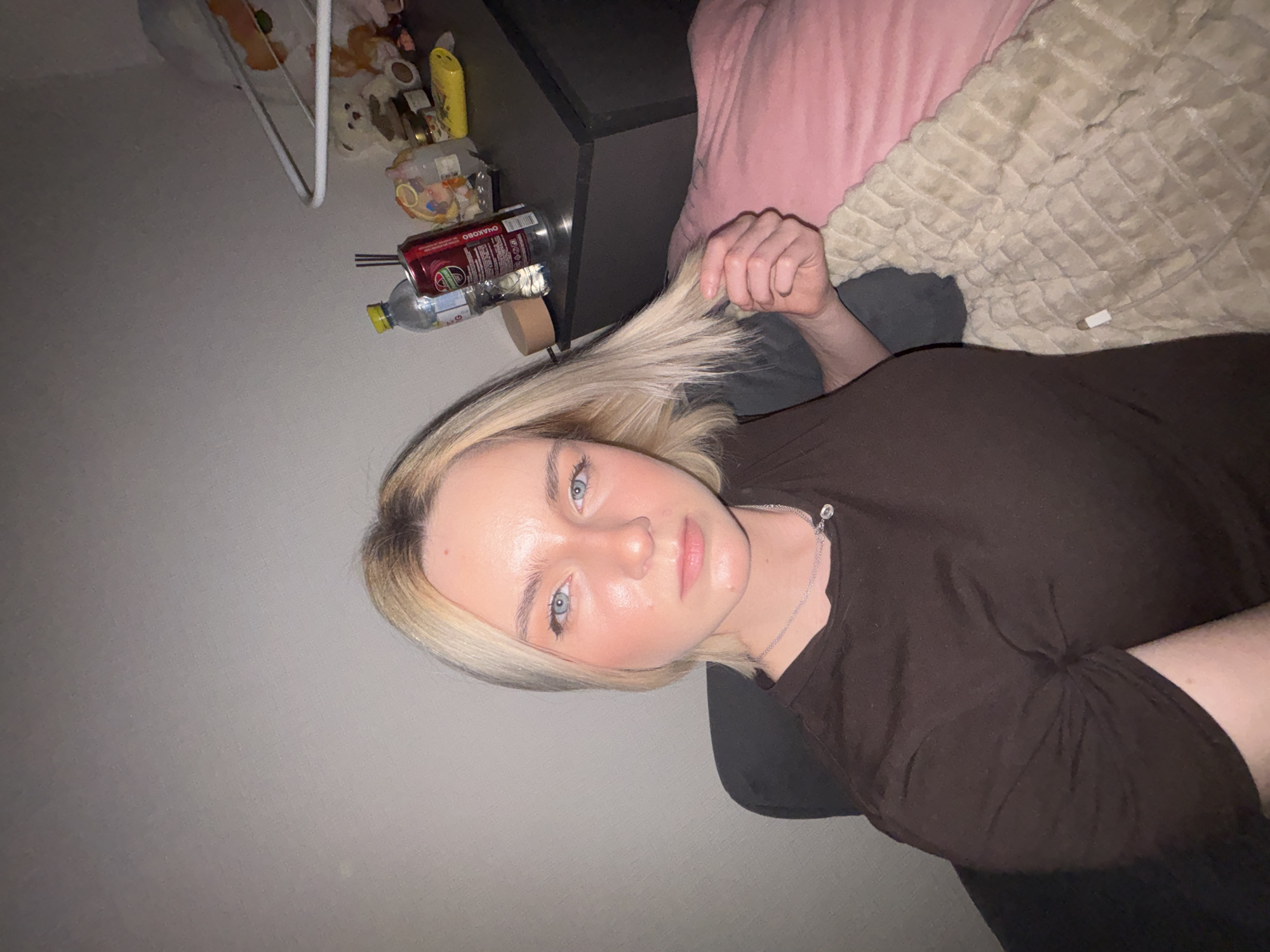 A young, fair-skinned woman with blue eyes and blonde hair is lying down, holding a strand of her hair. She wears a brown shirt and a silver necklace. She is partially covered by a pink pillow and a beige comforter. Beside her is a black bedside table with various items on it, including a can of "OHAKOBO," a water bottle, and a collection of small objects on a shelf above. The background includes a gray wall and a glimpse of the floor. The location information (Vsevolozhsk, Russia) is not verifiable from the image alone.