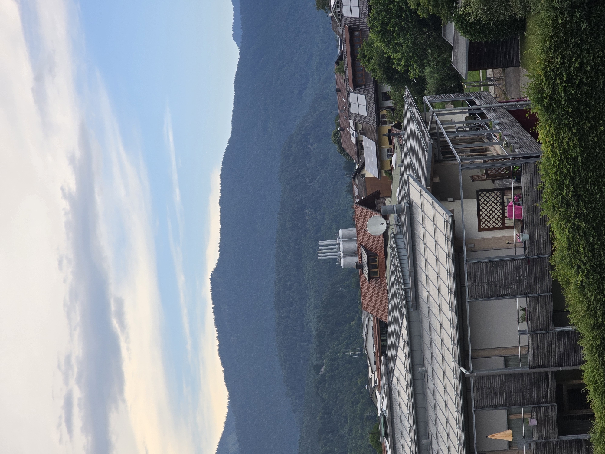 The image depicts a residential area with a backdrop of forested mountains and a hazy sky. In the foreground, a modern building with a metal roof and balconies is visible. The balconies feature wooden latticework screens and are adorned with potted plants and outdoor furniture, including a pink chair. Adjacent to this building is a smaller wooden shed. Further in the background, a cluster of houses with red-tiled roofs and solar panels can be seen. Dominating the mid-ground is a factory or industrial facility with several tall silver silos or tanks. The entire scene is framed by a lush green hedge in the immediate foreground and a dense, dark green forest covering the slopes of the mountains behind. The sky above is a mix of pale blue and diffused white and gray clouds. The image appears to be taken from an elevated viewpoint, suggesting it might be from another building or a hillside overlooking the town. This scene is consistent with the outskirts of Garmisch-Partenkirchen, Germany, given the mountainous terrain and architectural style of the buildings.