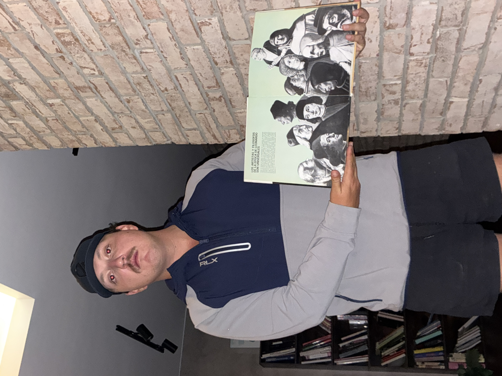 A man wearing a black cap, a blue and grey RLX jacket, and black shorts is holding open a book. The book has black and white illustrations of people and text in what appears to be Spanish. The man is standing in a room with a brick wall, grey walls, and a bookshelf filled with books. The location is Huixquilucan de Degollado, Mexico.