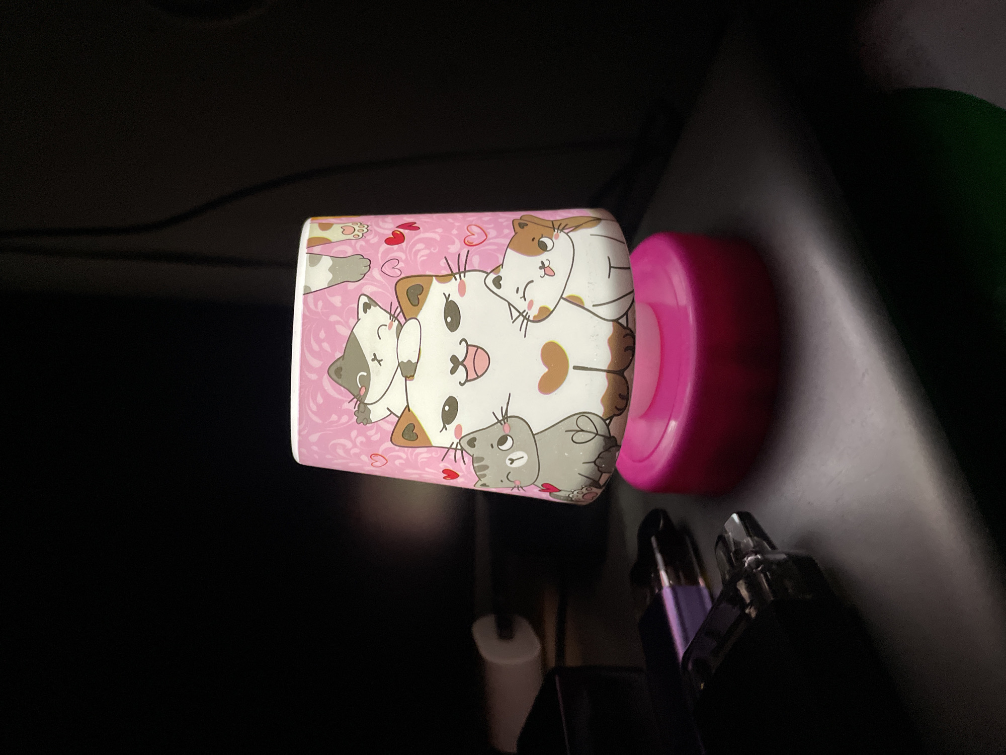 The image shows a small, pink night light decorated with cartoon cats sitting on a dark surface, possibly a shelf or table. The night light has a conical shade with a pink background featuring a repeating pattern of swirls and small red hearts. The cats are depicted in various poses and colors, with big eyes and happy expressions. One large white cat with brown markings is at the front, and smaller cats are stacked behind and beside it. The base of the night light is a solid, glossy pink. To the left of the night light, several dark objects are visible, including what appears to be electronic devices, possibly lighters or vape pens, and a white cylindrical object. The background is dark and out of focus, suggesting an indoor, dimly lit environment. There is no discernible location information related to Saint Petersburg, Russia.