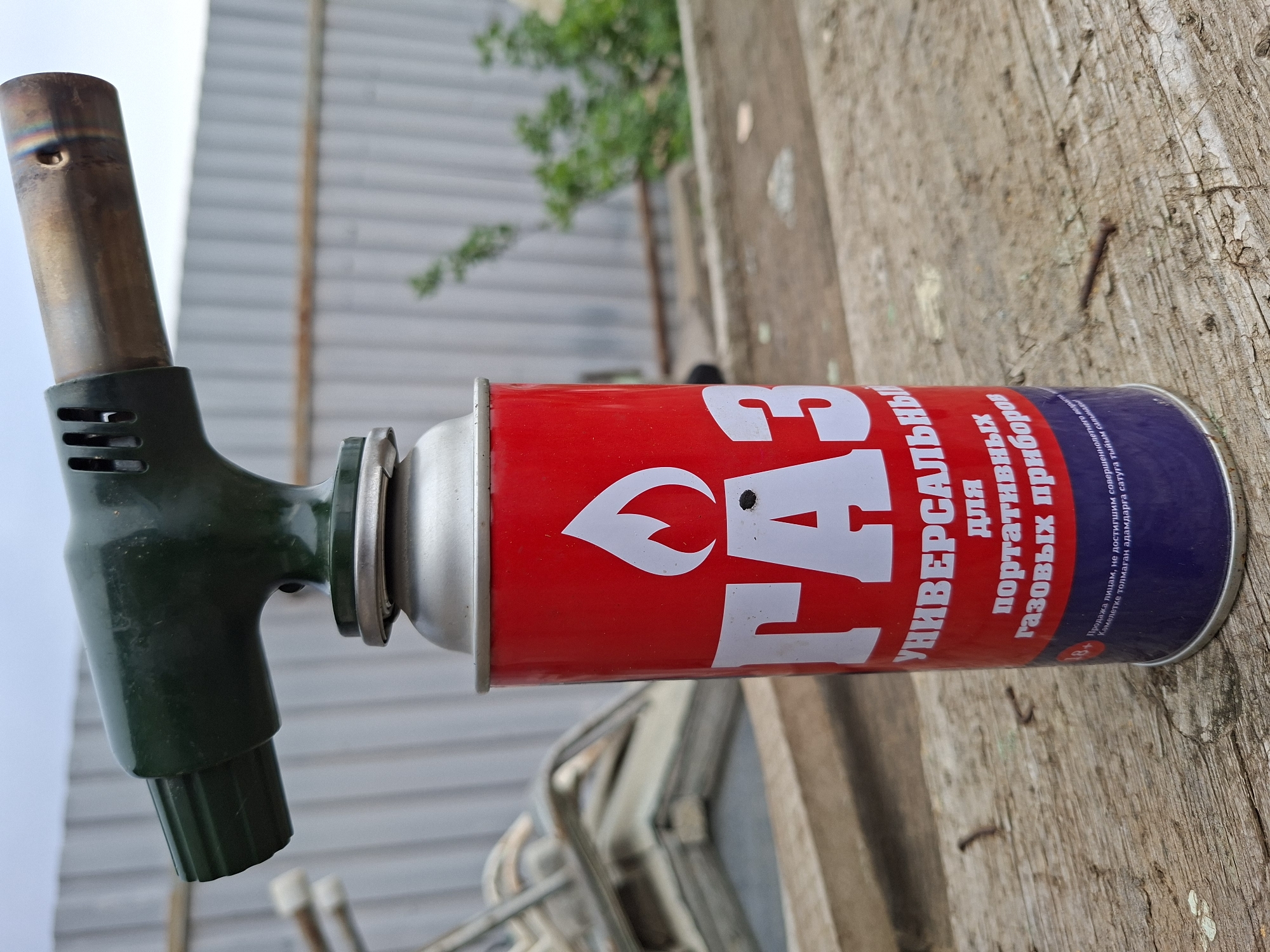A gas blowtorch is positioned on a weathered wooden surface outdoors. The blowtorch features a red canister with white Cyrillic lettering, attached to a green nozzle and a metal tip. The lettering includes the word "GAZ" in large, bold font. The background reveals a corrugated metal fence, some greenery, and a glimpse of the sky.
