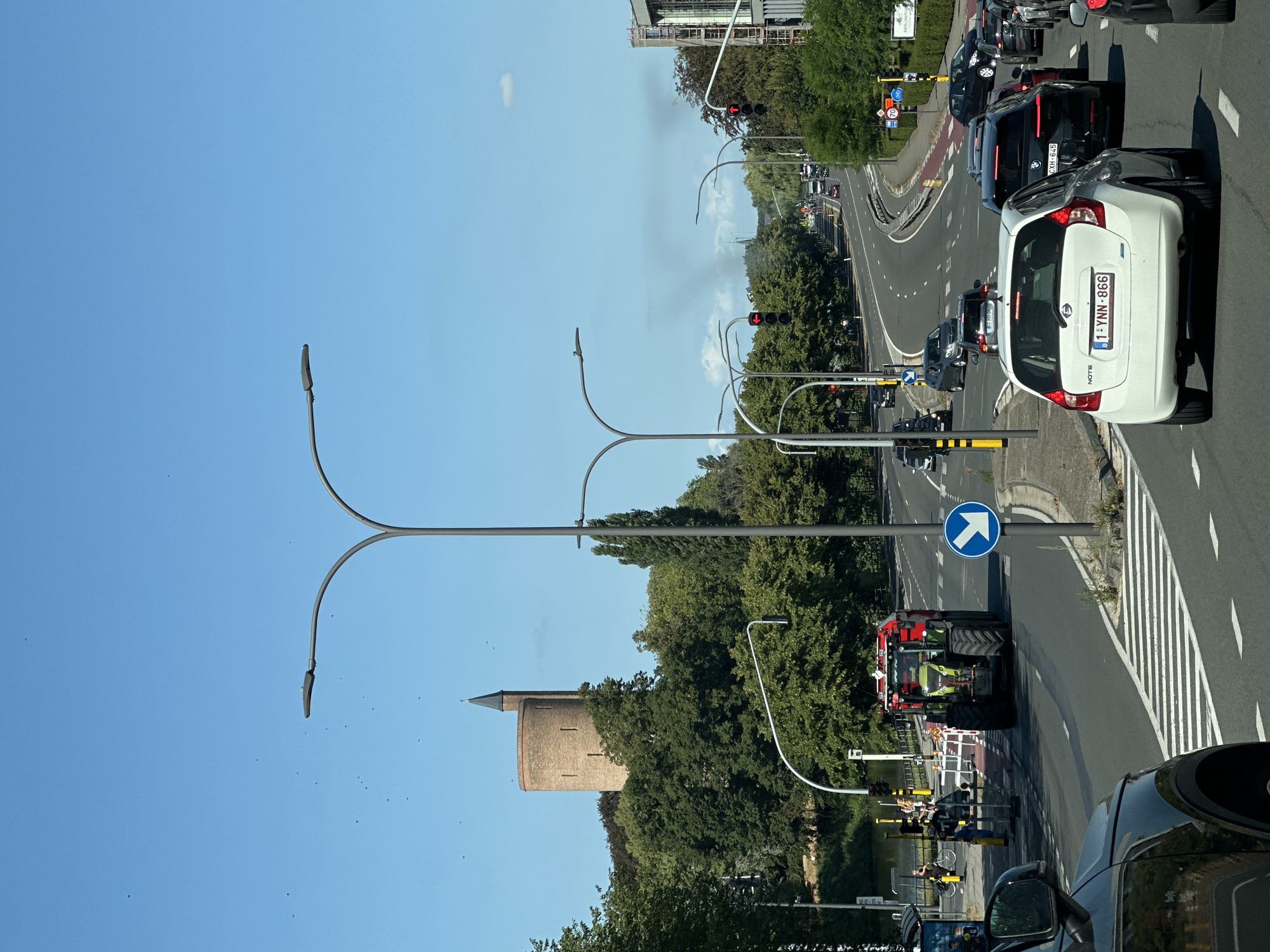 The image shows a sunny day in Bruges, Belgium, with a busy road scene. In the distance, a historic brick tower stands amidst lush green trees. The foreground features a traffic intersection with several cars, including a white hatchback with a Belgian license plate "1-YNN-866", and a red tractor with a trailer. Traffic lights are visible, and a blue directional arrow sign is posted on a pole. People are seen near the roadside, with some individuals on bicycles. The environment is urban, with trees lining the road and buildings in the background. Smoke is rising in the distance, obscuring some of the view.