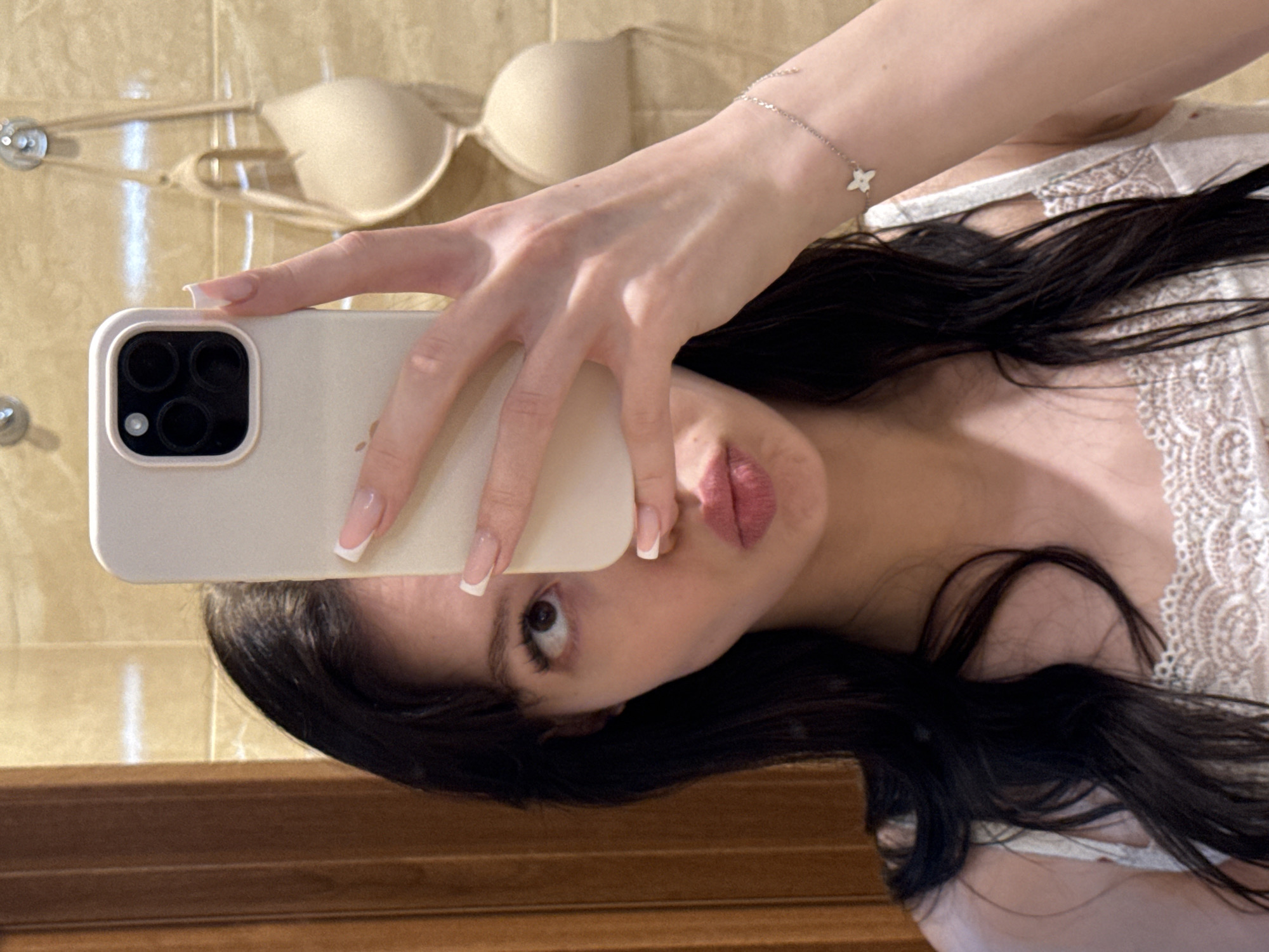 A young woman is taking a selfie in a bathroom. She is holding a smartphone in her right hand, obscuring the left side of her face. Her left eye is visible, looking upwards, and her lips are pursed. She has long, dark hair and is wearing a white top with lace detailing. A beige bra is hanging on the wall in the background. The bathroom appears to have beige tiled walls and a wooden door frame. The image does not contain any information that could be used to determine the location in the city of Khimki, Russia.