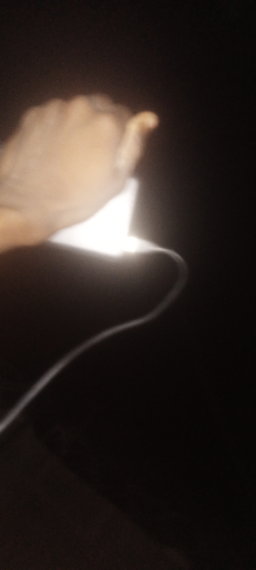 A photograph of a hand holding a brightly illuminated object with an attached cord against a very dark background. The hand is in the upper left portion of the image, and the cord extends in a curving line toward the lower left. It is not possible to discern specific details about Sabon Tasha, Nigeria from the image alone.