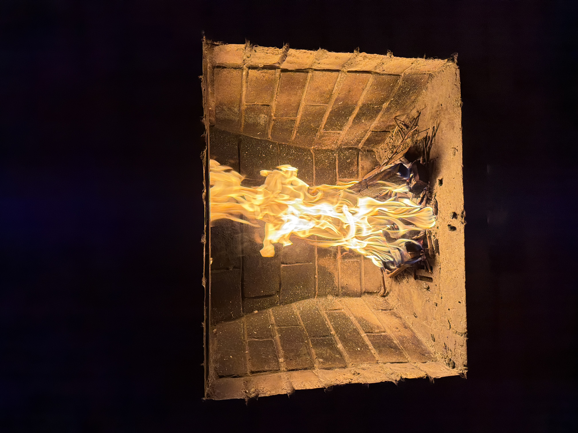 An indoor fireplace is depicted with a fire burning brightly within its brick-lined cavity. The flames are a vibrant orange and yellow, casting a warm glow on the surrounding brickwork and the ash-covered hearth. The fireplace appears to be made of rustic brick, with some rough edges and a slightly worn appearance. The background is dark and indistinct, suggesting an evening or nighttime setting, or a dimly lit room. There is no indication of the specific location in Қарғалы, Kazakhstan. There are no people visible in the image, and the focus is solely on the fireplace and the fire.