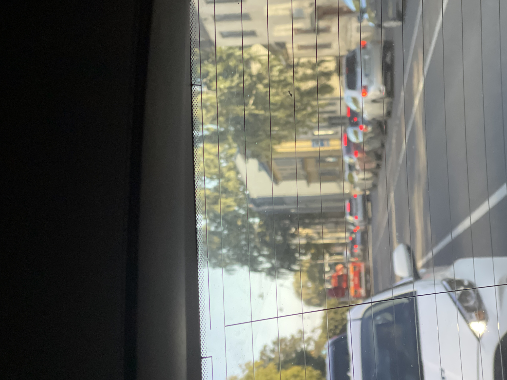 The image shows a view through a car's rear window, looking out onto a street in Tallinn, Estonia, during daylight hours. The scene depicts a line of traffic, suggesting a possible traffic jam or slow-moving traffic. Several cars are visible, including a white car in the foreground. In the distance, a red truck is present. Buildings line the street, with trees providing greenery. The sun is shining, casting light and shadows. The perspective is from inside a vehicle, looking outwards, with horizontal lines from the rear window defroster elements running across the image.