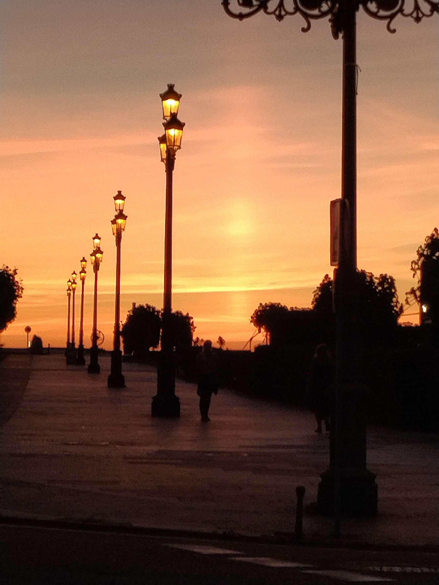 This image depicts an outdoor scene at dusk, likely in Baiona, Spain, characterized by a row of lampposts lining a paved walkway. The sky is a vibrant gradient of orange and pink, indicating sunset or sunrise. The lampposts, illuminated with a warm glow, are spaced along the path, casting light on the ground. Silhouetted against the sky and the ambient light are several individuals walking along the path. Trees and foliage are visible in the background, also rendered as dark silhouettes. The overall impression is one of a tranquil evening or early morning stroll in a public area.