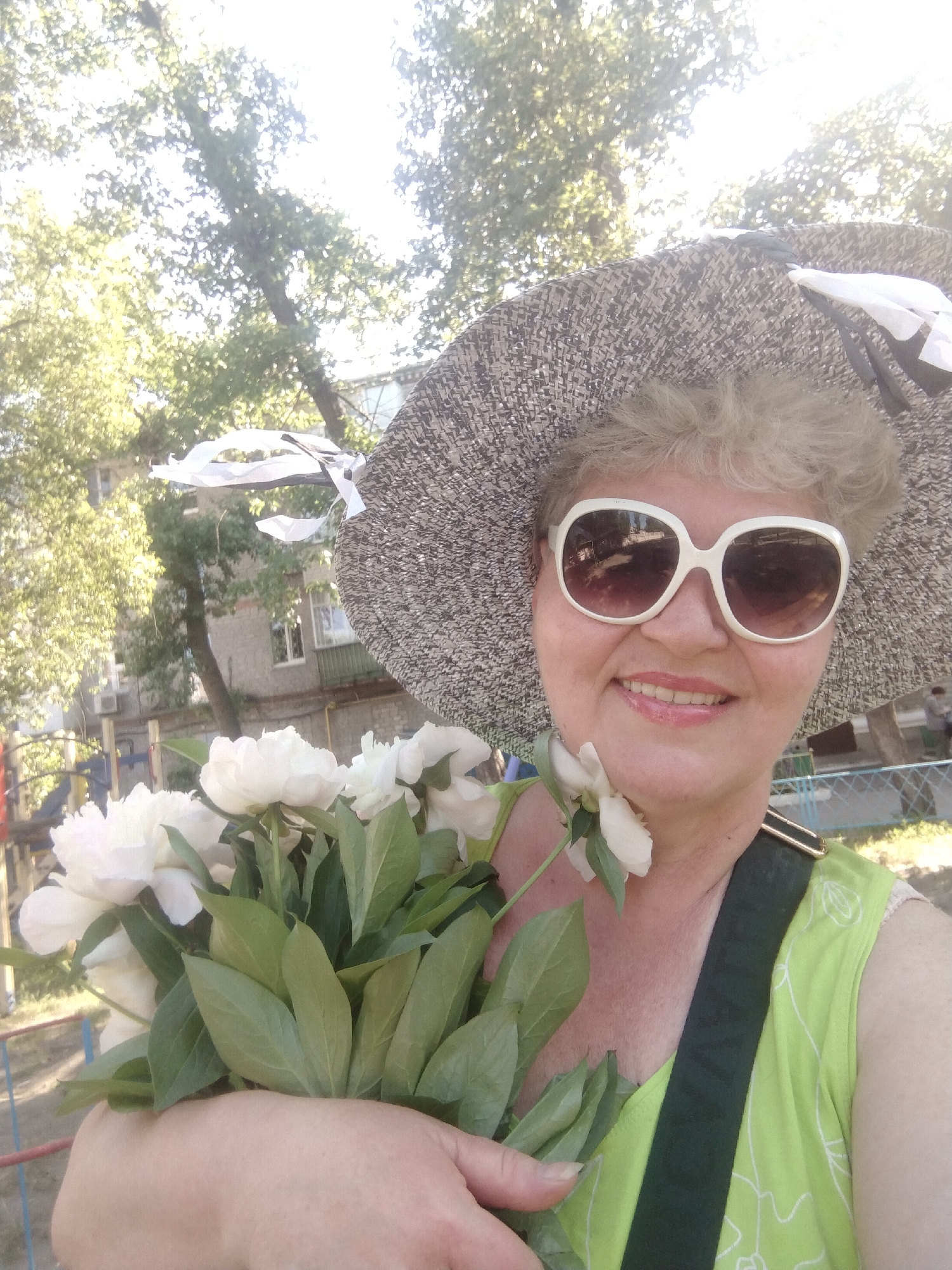 The image is a close-up selfie of a woman holding a bouquet of white peonies in an outdoor setting. The woman is wearing a wide-brimmed straw hat with white ribbons and large, white-rimmed sunglasses. She has blonde, permed hair and is smiling. She is wearing a light green, sleeveless top with a white leafy pattern and a dark green strap across her chest. The background suggests a city environment in Horishni Plavni, Ukraine, with trees, buildings, and a playground visible. The lighting indicates it is daytime, likely sunny.